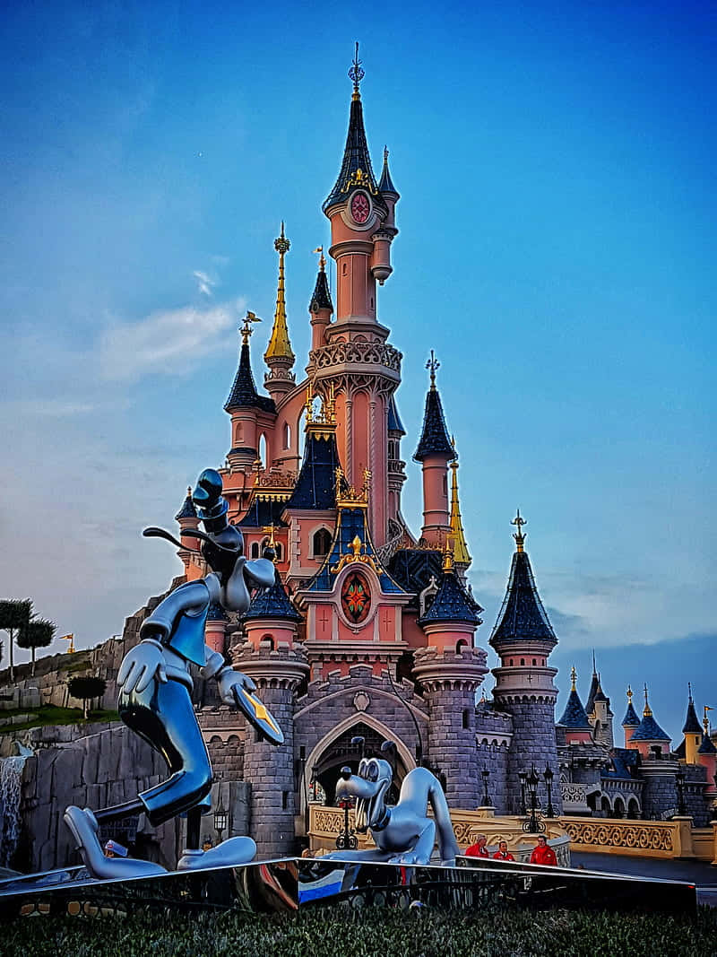 Disneyland Castle 桌布 壁紙 — 一座宏偉的粉紅色城堡，擁有藍色屋頂和金色尖塔，在晴朗的藍天下高聳矗立，兩旁是大型金屬迪士尼角色雕像。