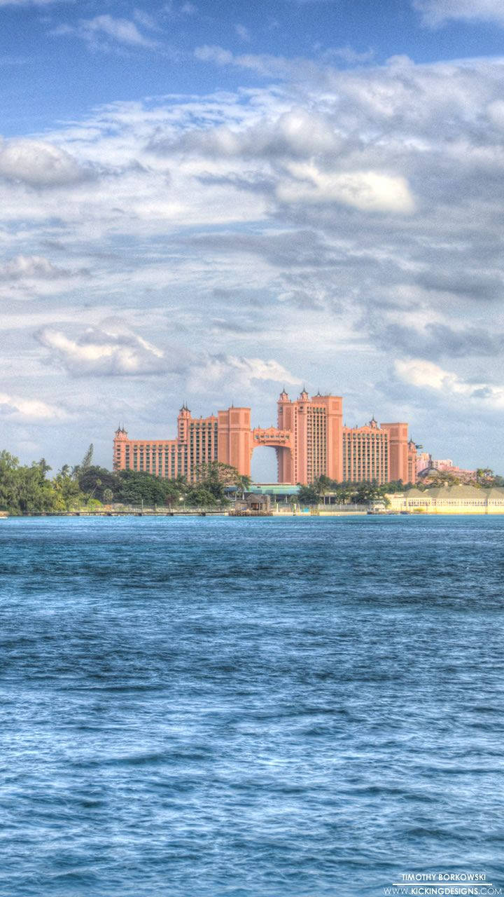 Nassau Bahamas 桌布 壁紙 — 一座宏偉的粉紅色度假酒店從一個鬱鬱蔥蔥的島嶼上聳立而起，越過閃爍的藍色水域，天空部分多雲。