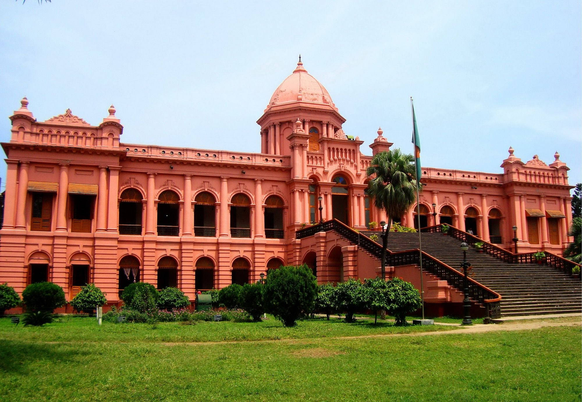 Dhaka 桌布 壁紙 — 一座宏偉的粉紅色歷史建築，擁有圓頂屋頂和拱形窗戶，在晴朗的藍天下雄偉地矗立在修剪整齊的草坪上。