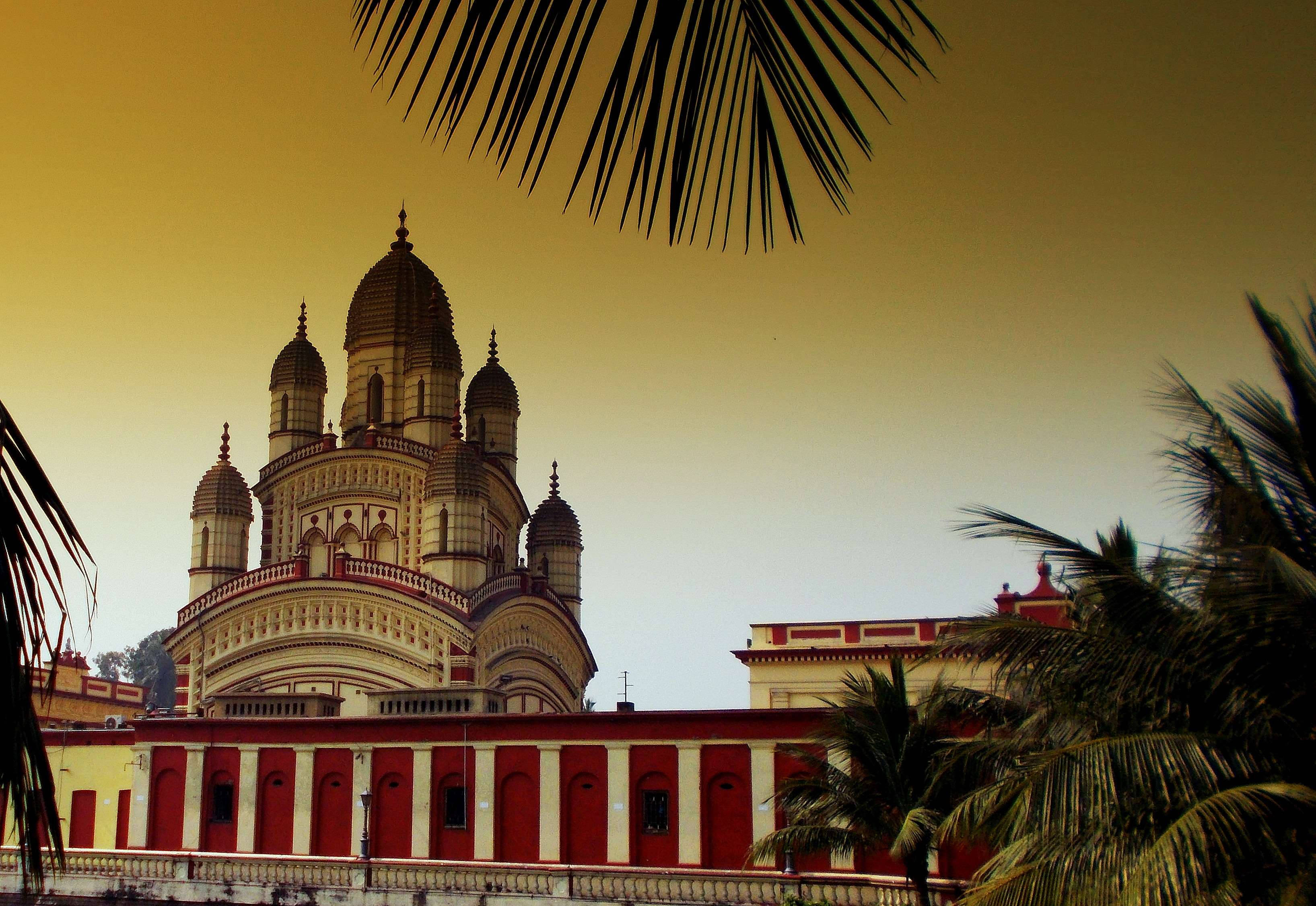 Kolkata 桌布 壁紙 — 一座宏偉的紅白相間圓頂寺廟，在金色夕陽天空下，聳立在棕櫚葉的剪影之後。