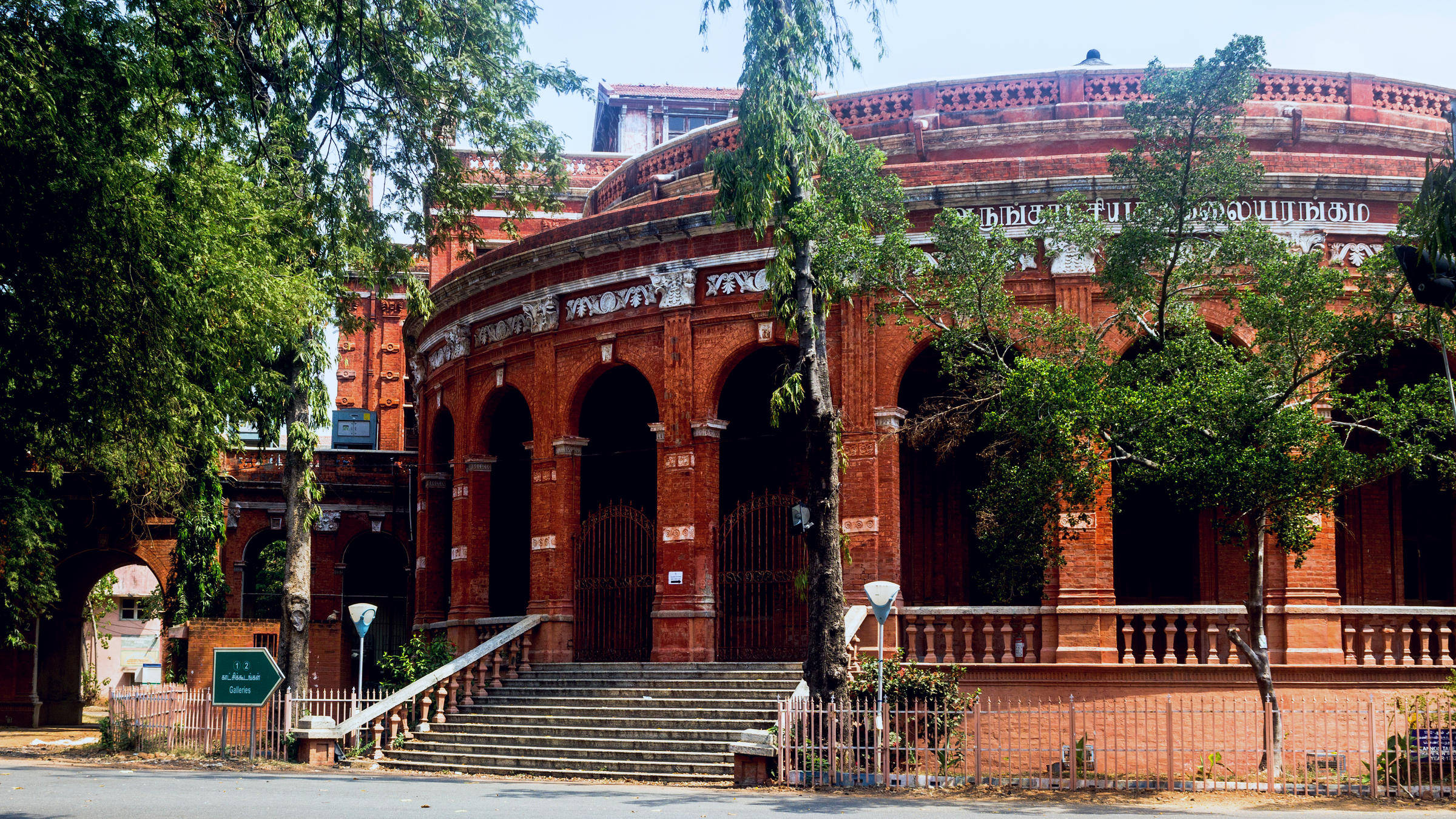 Chennai 桌布 壁紙 — 一座宏偉的紅磚圓形建築，擁有拱形入口和華麗雕刻，在明媚陽光下矗立，四周環繞著茂密的綠樹。
