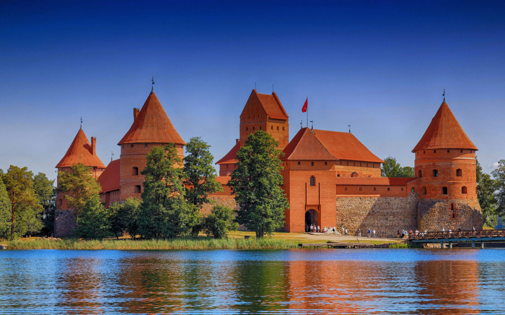 Vilnius 桌布 壁紙 — 一座宏偉的紅磚城堡，帶有圓錐形塔樓，矗立在平靜的湖邊，天空湛藍。