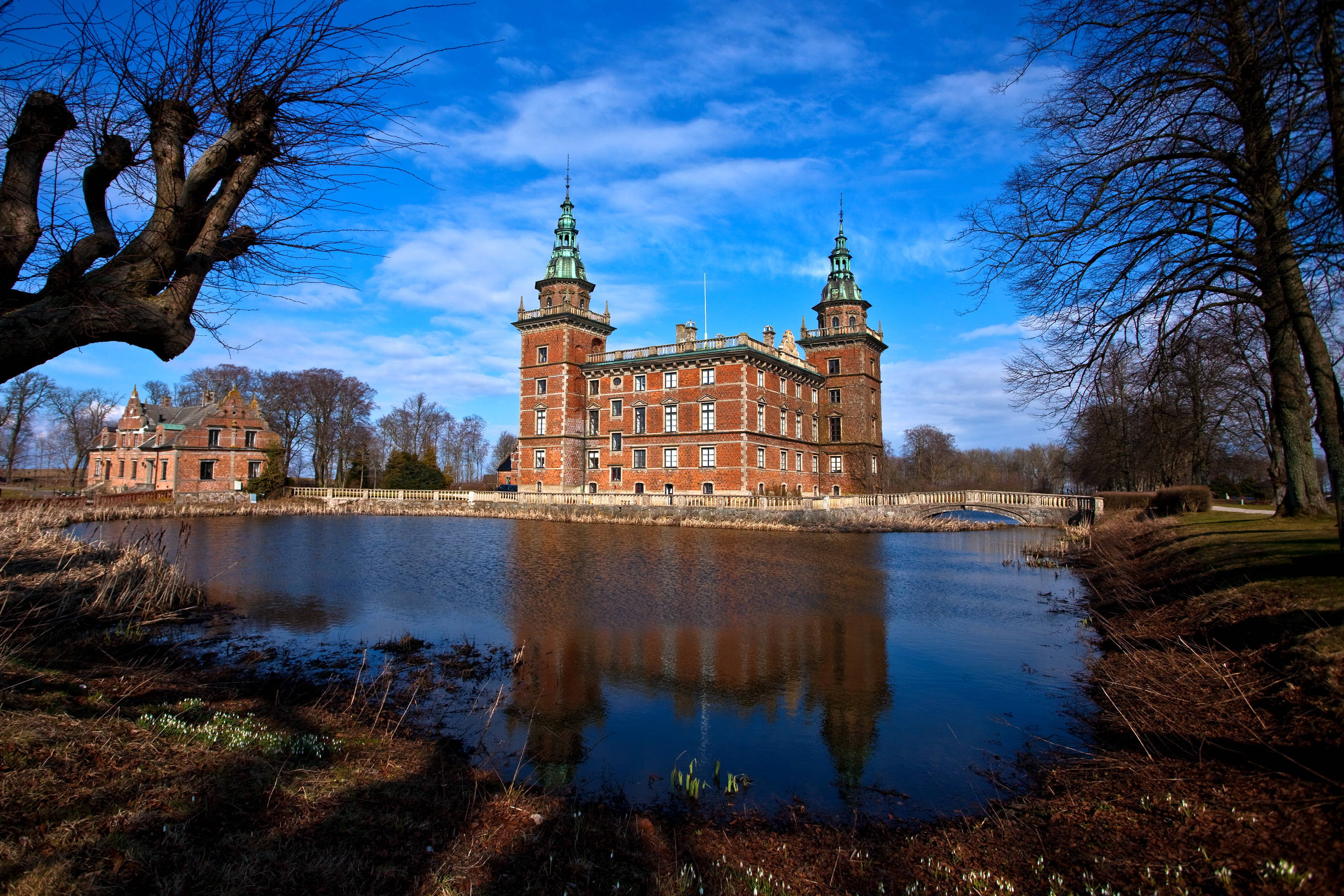 Sweden 桌布 壁紙 — 一座宏偉的紅磚城堡，帶有綠色尖頂，倒映在平靜的池塘中，上方是晴朗的藍天，點綴著幾朵白雲。