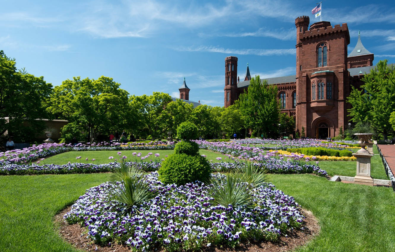 Smithsonian 桌布 壁紙 — 一座宏偉的紅磚城堡式建築矗立在充滿活力的花壇和修剪整齊的草坪後面，頭頂是清澈的藍天。