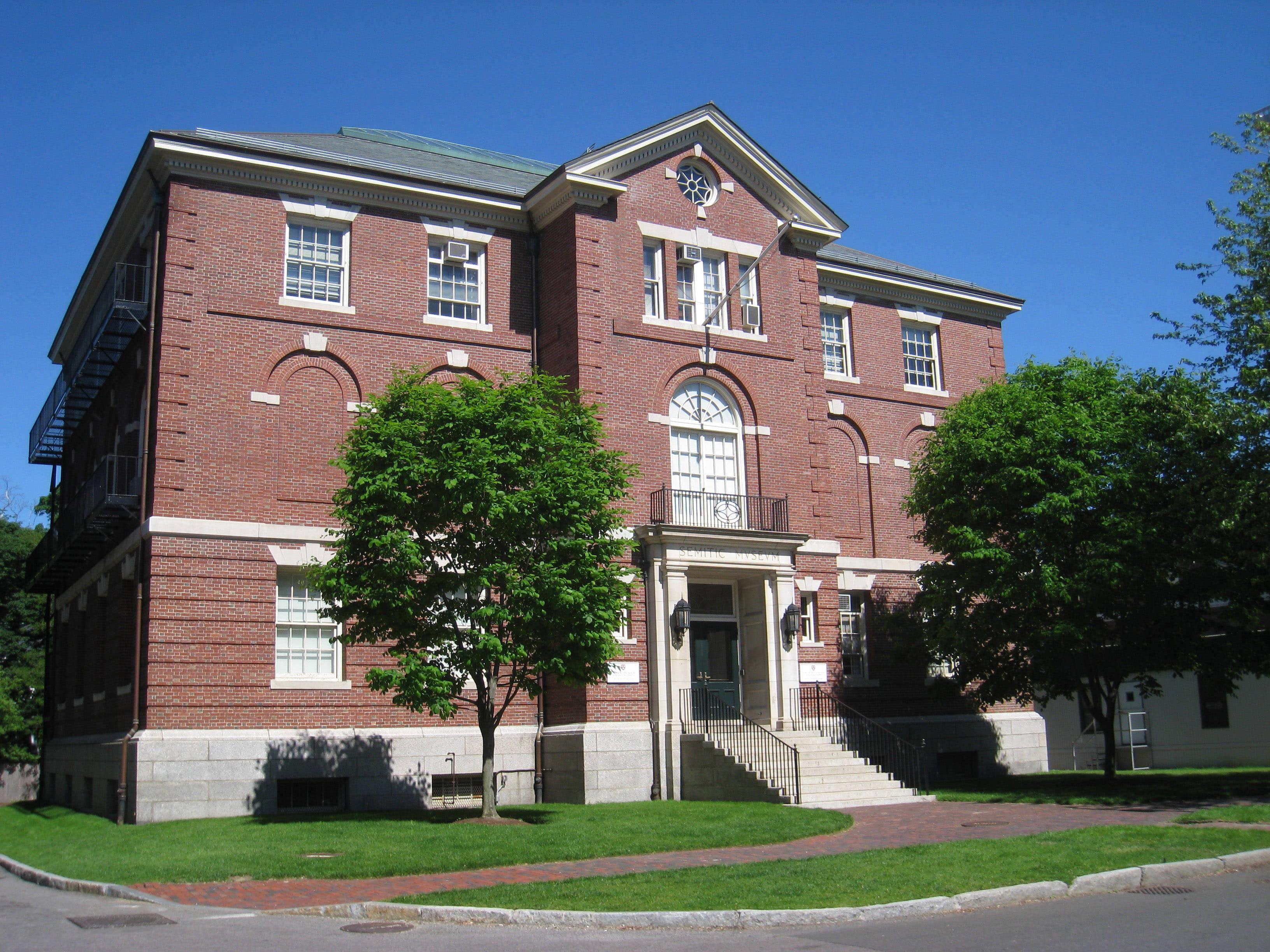Museum 桌布 壁紙 — 一座宏偉的紅磚大學建築，帶有白色飾邊和拱形窗戶，矗立在晴朗的藍天下，周圍環繞著茂密的綠樹。