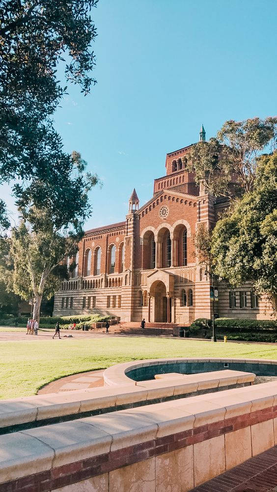 Library 桌布 壁紙 — 一座宏偉的紅磚大學建築，擁有拱形窗戶和中央塔樓，矗立在晴朗的藍天下，周圍環繞著茂密的綠樹和修剪整齊的草坪，前景有彎曲的水景。