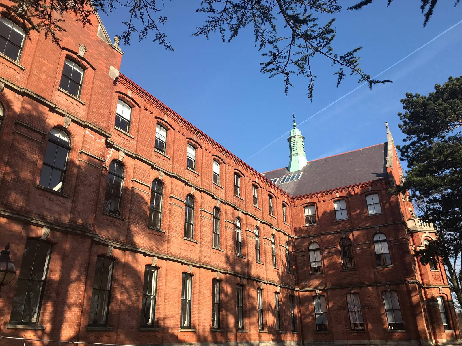 桌布 壁紙 — 一座宏偉的紅磚大學建築，擁有拱形窗戶和綠色圓頂，矗立在樹枝框住的清澈藍天下。