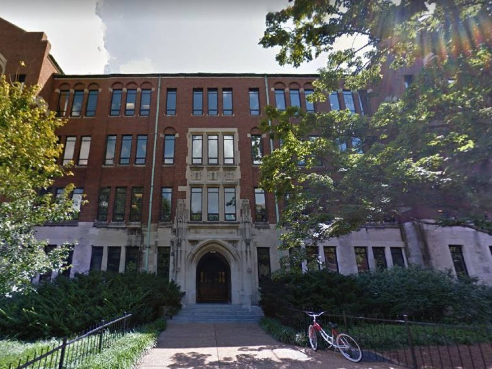 Building 桌布 壁紙 — 一座宏偉的紅磚大學建築，擁有拱形窗戶和石頭入口，周圍環繞著茂密的綠樹和一輛停放的自行車。