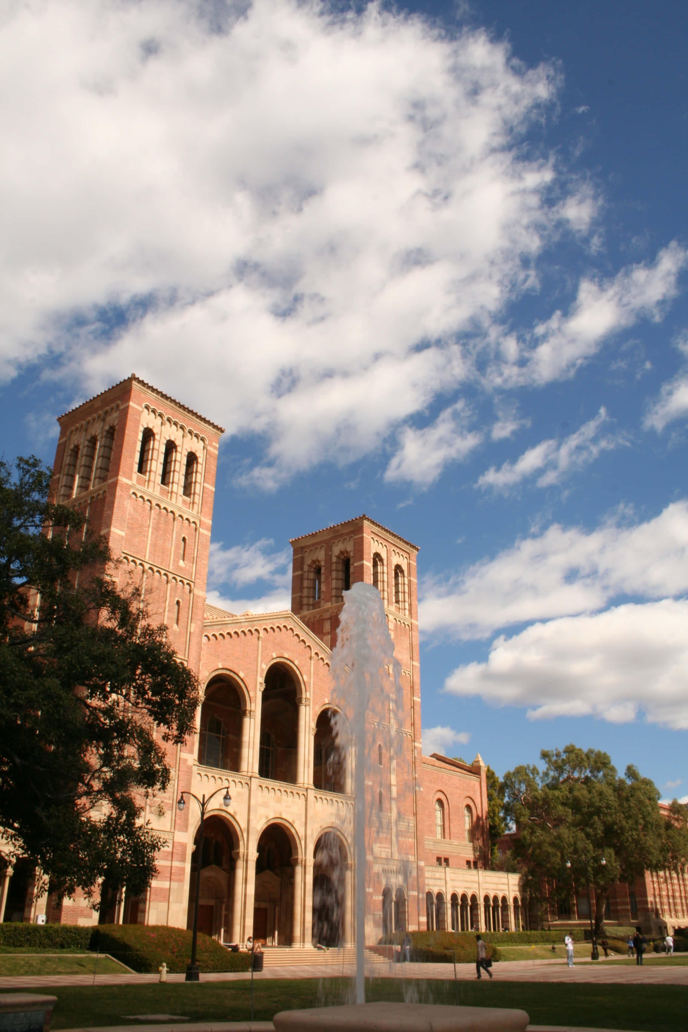 Ucla Campus 桌布 壁紙 — 一座宏偉的紅磚大學建築，擁有雙塔和拱形步道，矗立在晴朗的藍天下，點綴著蓬鬆的白雲，中央噴泉和綠色草坪環繞其間。