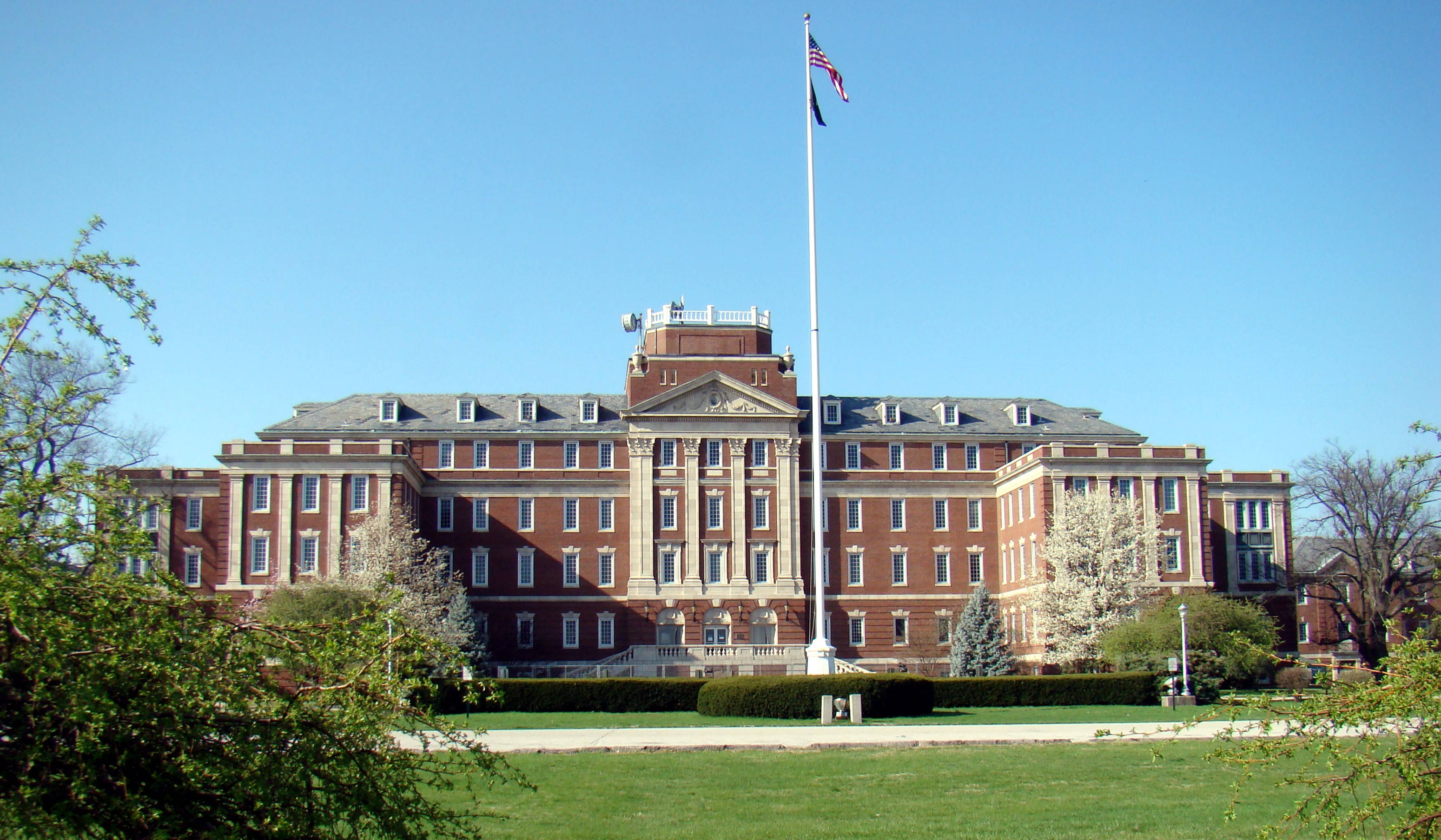 Hospital 桌布 壁紙 — 一座宏偉的紅磚大學建築，配有白色柱子和旗桿，矗立在晴朗的藍天下，周圍環繞著綠色草坪和萌芽的樹木。