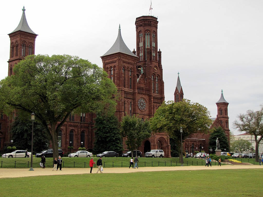 Smithsonian 桌布 壁紙 — 一座宏偉的紅磚歷史建築，擁有多座塔樓和大型鐘面，矗立在茂密的綠樹和草地公園之後，公園內有行人沿著小徑行走。