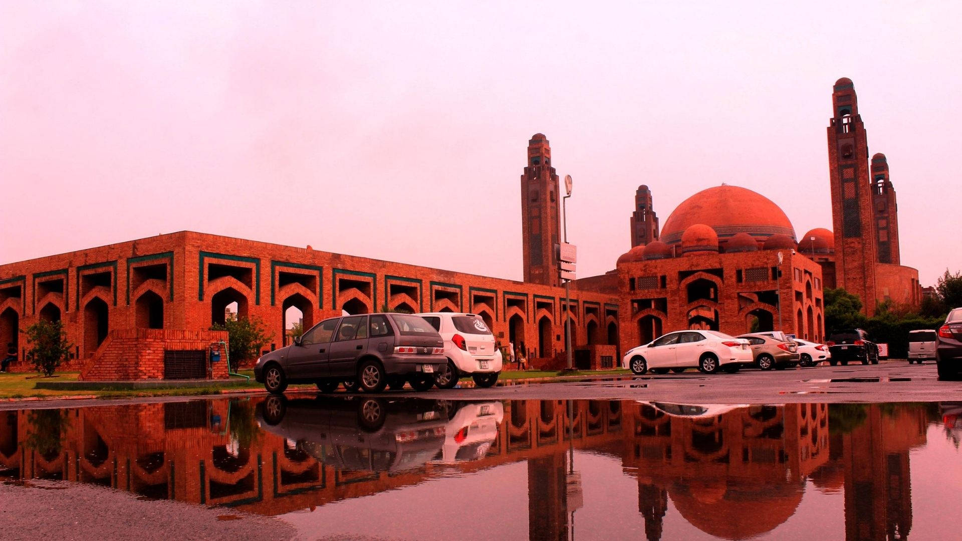 Pakistan 桌布 壁紙 — 一座宏偉的紅磚清真寺，擁有圓頂和宣禮塔，倒映在雨水中，附近有汽車停泊在粉紅色的天空下。