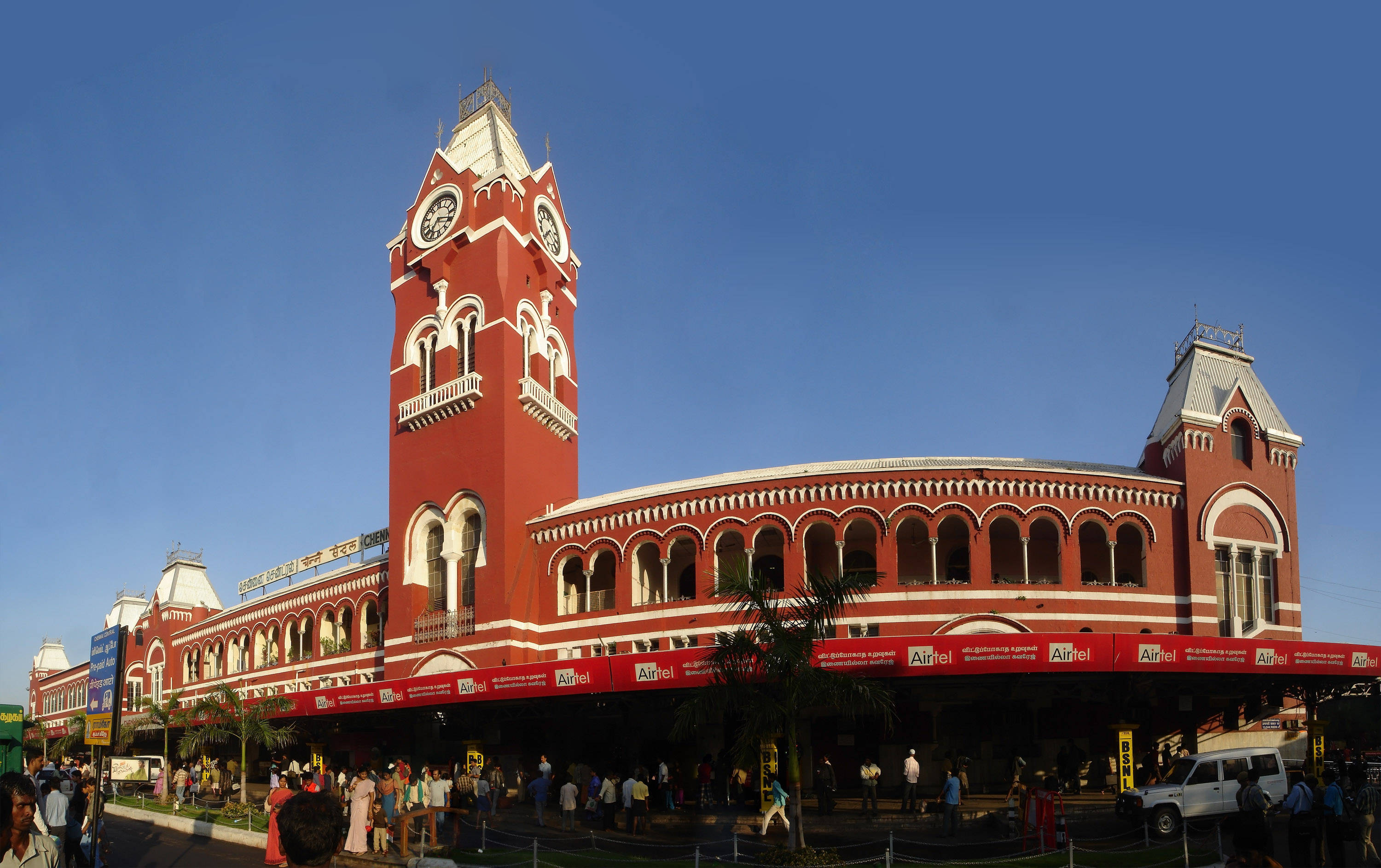 Chennai 桌布 壁紙 — 一座宏偉的紅磚火車站，高聳的鐘樓矗立在晴朗的藍天下，入口處聚集著人群。