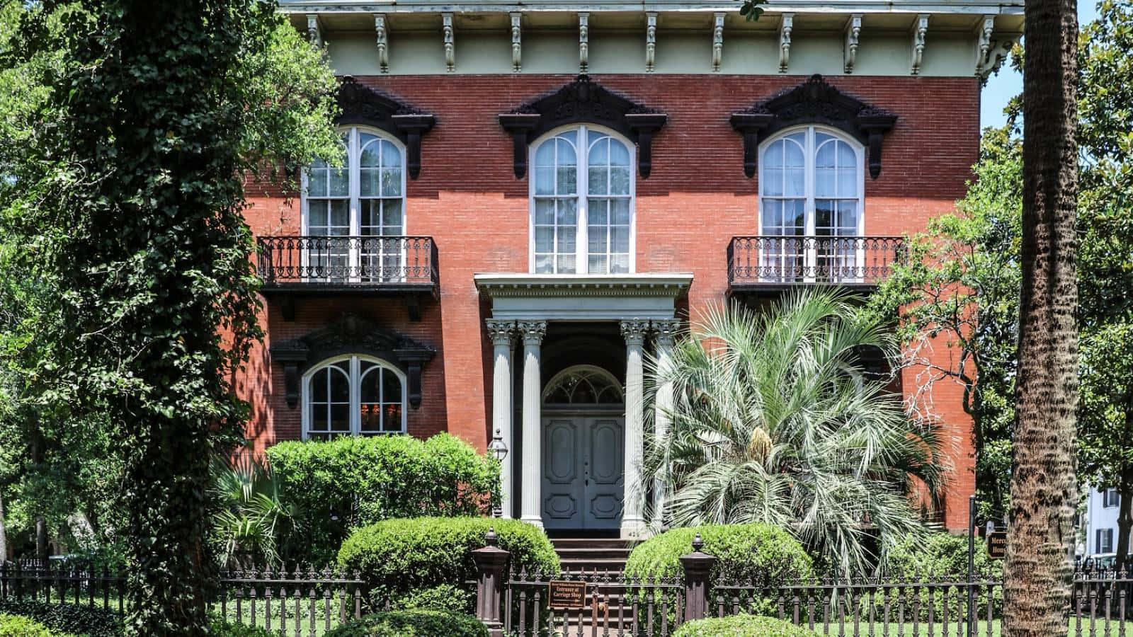 Savannah 桌布 壁紙 — 一座宏偉的紅磚維多利亞式房屋，帶有白色飾邊和拱形窗戶，在明亮的日光下被茂密的綠植和棕櫚樹環繞。