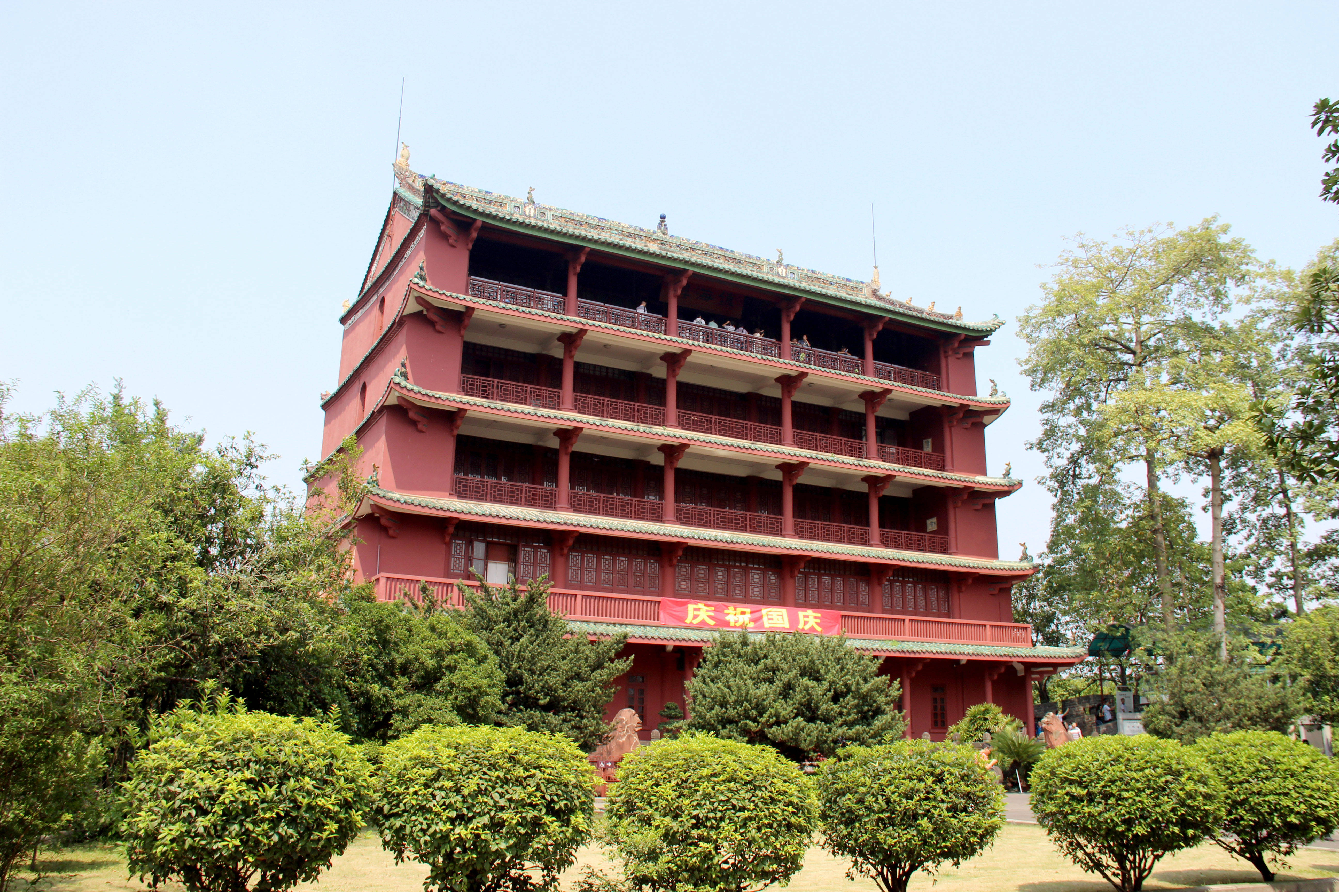 Guangzhou 桌布 壁紙 — 一座宏偉的紅色傳統中式寶塔，配以綠色瓦片屋頂，在晴朗的藍天下矗立於茂密的綠樹和修剪整齊的灌木叢中。