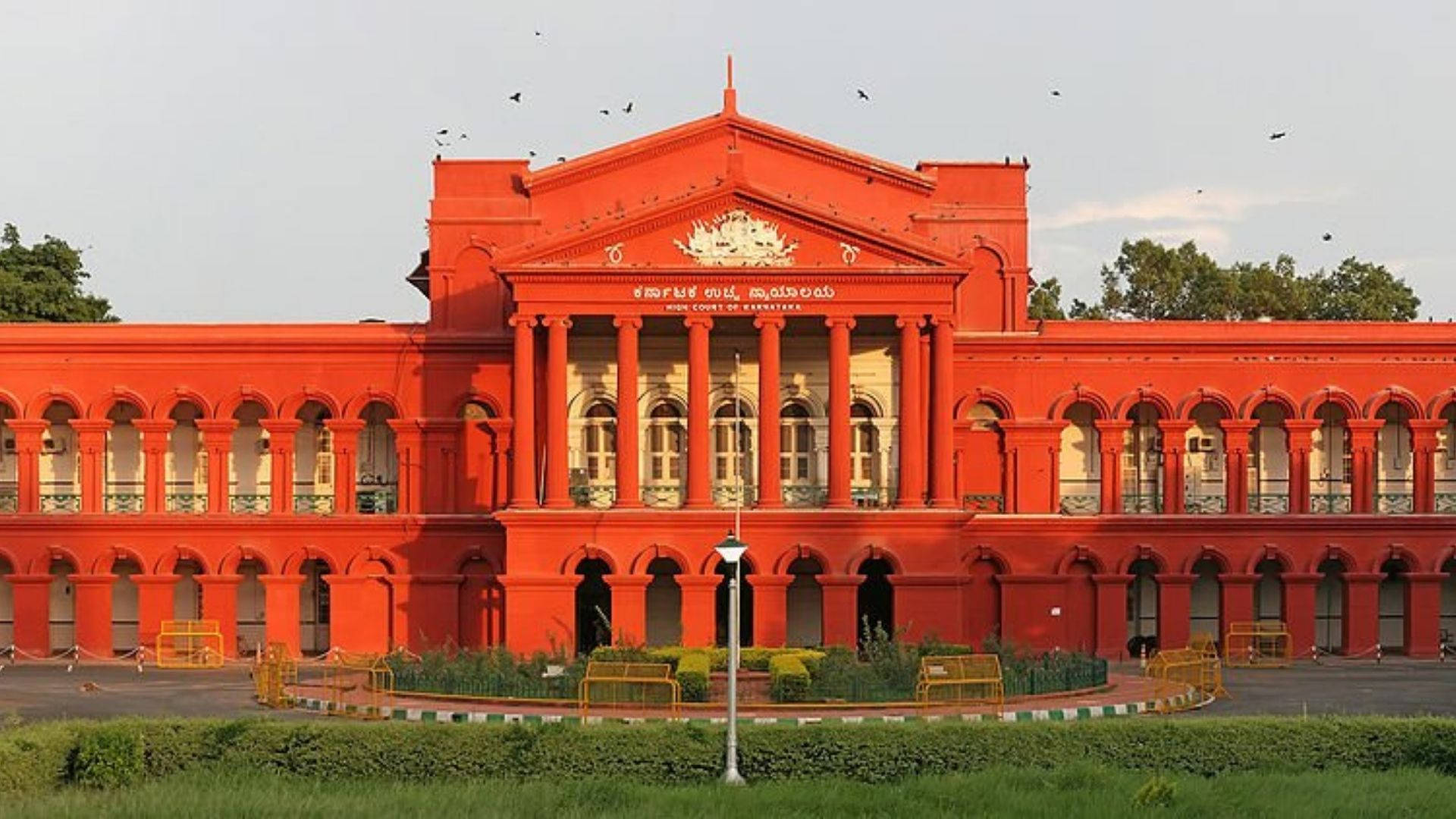 Bangalore 桌布 壁紙 — 一座宏偉的紅色砂岩法院大樓，擁有古典柱子和拱形窗戶，矗立在蒼白的天空下，鳥兒在上空飛翔。