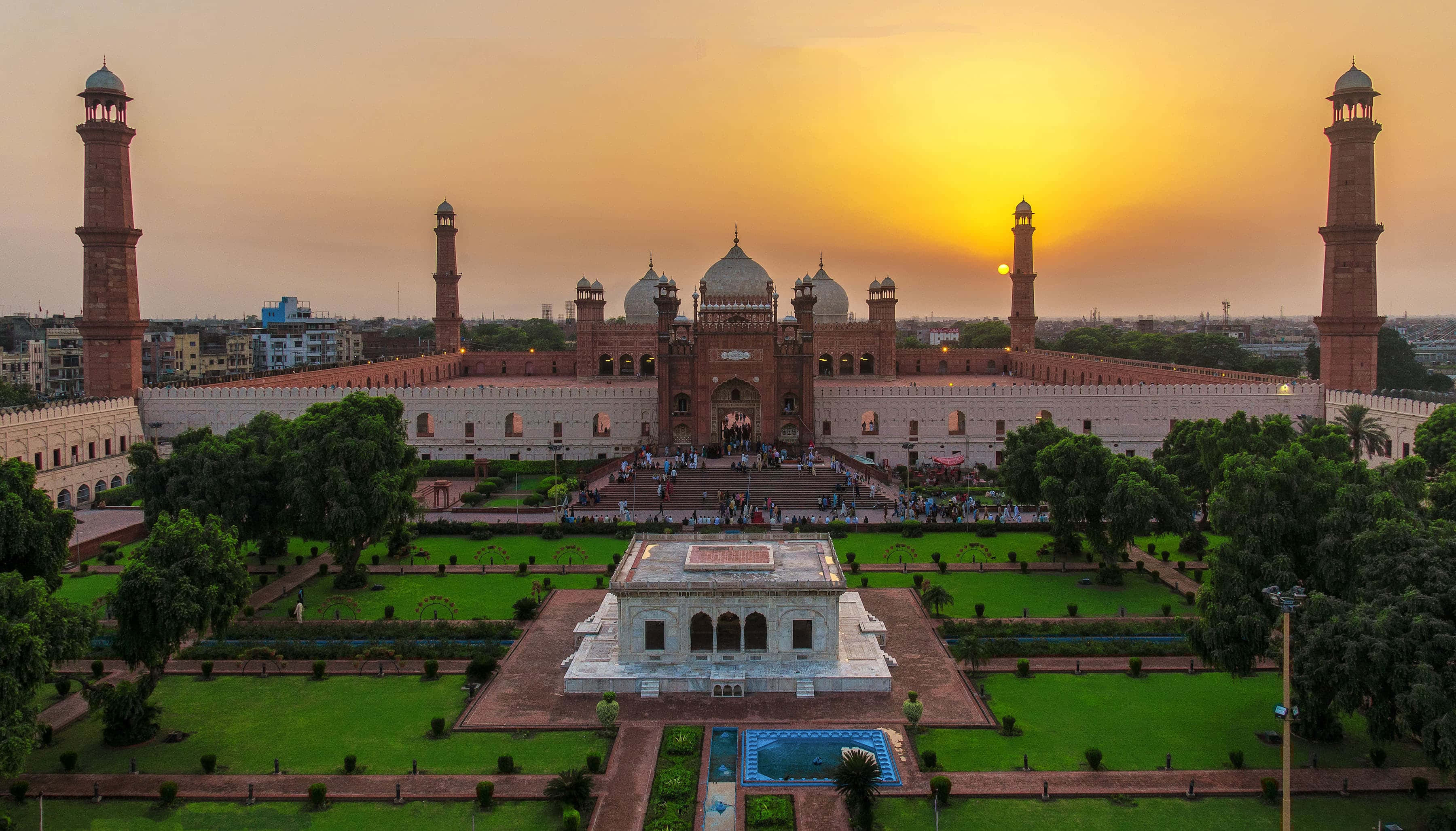 Pakistan 桌布 壁紙 — 一座宏偉的紅色砂岩清真寺，擁有四座宣禮塔，沐浴在金色的日落光芒中，周圍環繞著鬱鬱蔥蔥的花園。