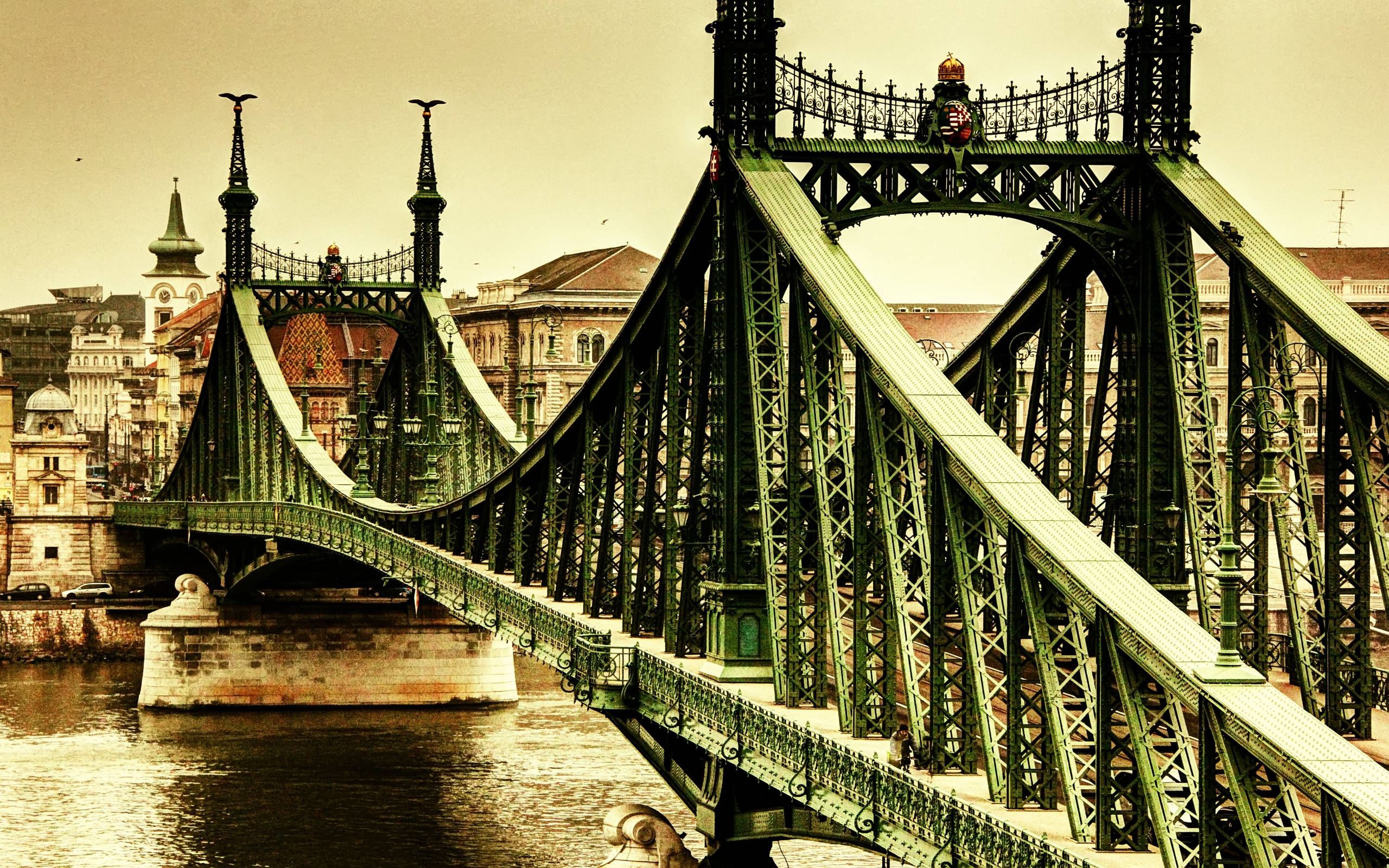 Hungary 桌布 壁紙 — 一座宏偉的綠色鍛鐵橋拱跨在河上，河岸兩旁歷史建築林立，天空呈現溫暖的懷舊色調。