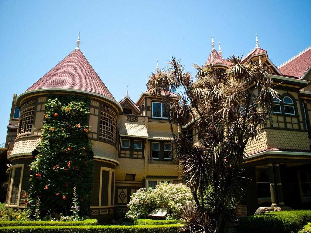 Mansion 桌布 壁紙 — 一座宏偉的維多利亞式大宅，擁有紅色瓦片屋頂和華麗的木工，矗立在晴朗的藍天下，周圍環繞著茂密的綠植和棕櫚樹。