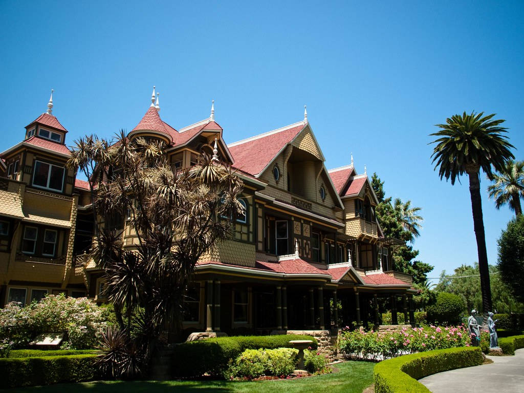 Mansion 桌布 壁紙 — 一座宏偉的維多利亞式大宅，擁有紅色瓦片屋頂和華麗的木工裝飾，矗立在晴朗的藍天下，周圍環繞著棕櫚樹和修剪整齊的花園。