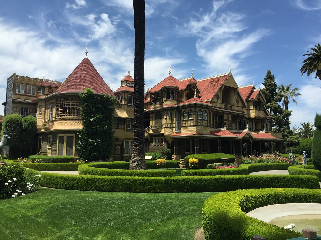 Mansion 桌布 壁紙 — 一座宏偉的維多利亞式大宅，擁有紅色瓦片屋頂和華麗的木工，矗立在修剪整齊的綠色草坪和棕櫚樹之間，在明亮的藍天下。