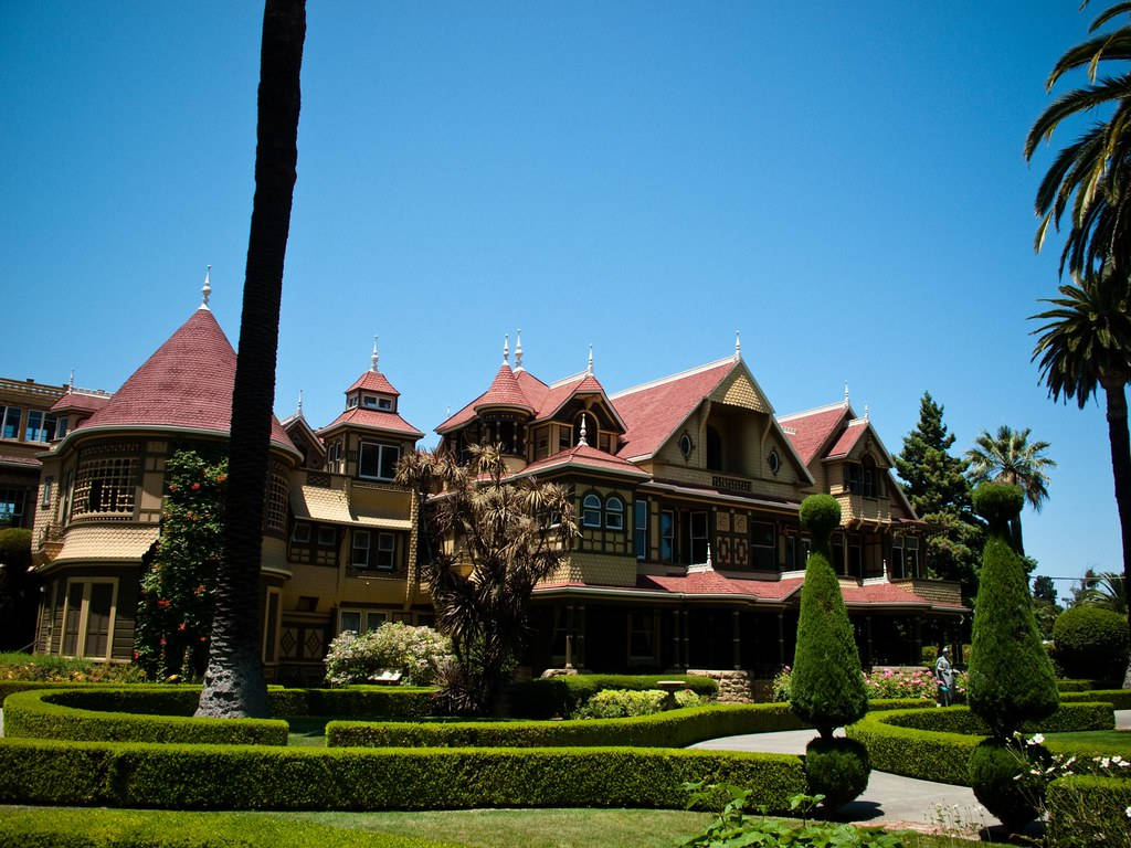 Mansion 桌布 壁紙 — 一座宏偉的維多利亞式大宅，擁有紅色瓦片屋頂和華麗的木工裝飾，矗立在晴朗的藍天下，周圍環繞著修剪整齊的樹籬和棕櫚樹。