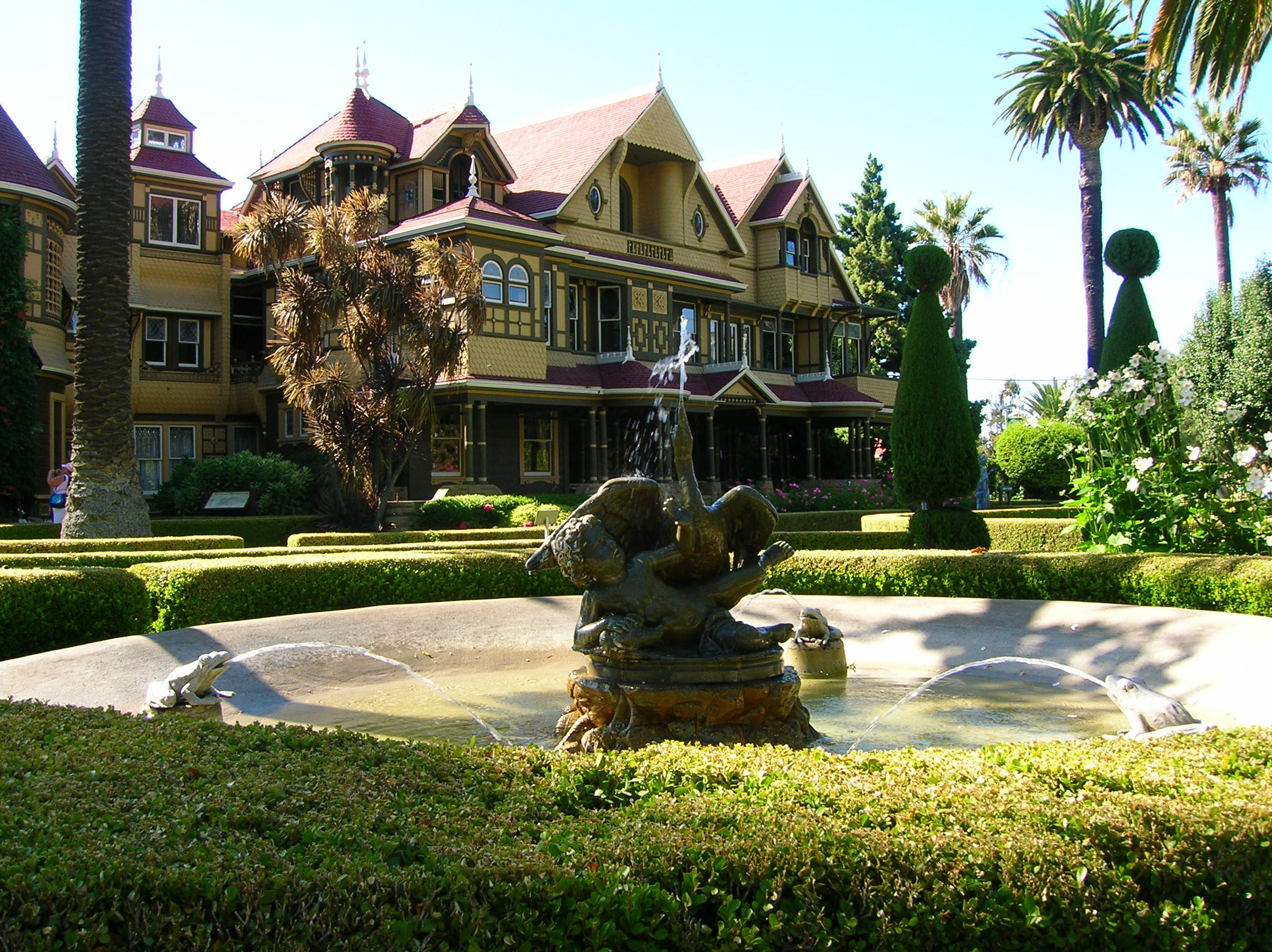 Mansion 桌布 壁紙 — 一座宏偉的維多利亞式大宅，擁有紅色屋頂和華麗的裝飾線條，矗立在噴泉後方，噴泉有雕塑人物和修剪整齊的樹籬，在明媚的陽光下。