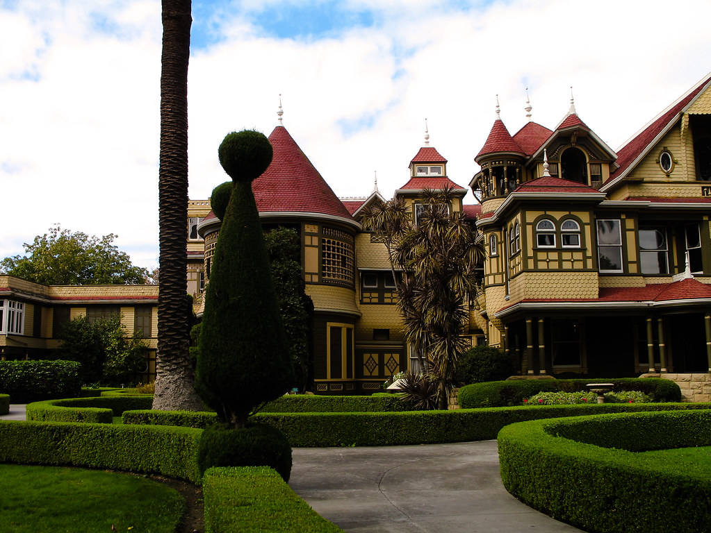 Mansion 桌布 壁紙 — 一座宏偉的維多利亞式豪宅，擁有紅色瓦片屋頂和華麗的木製品，矗立在修剪整齊的樹籬和棕櫚樹之間，天空部分多雲。