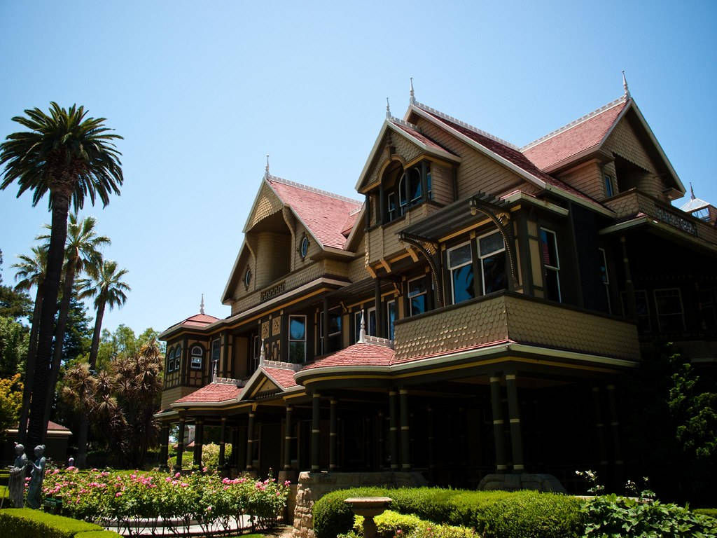 Mansion 桌布 壁紙 — 一座宏偉的維多利亞式豪宅，擁有紅色瓦屋頂和華麗的木製品，矗立在晴朗的藍天下，周圍環繞著棕櫚樹和盛開的粉紅色花朵。