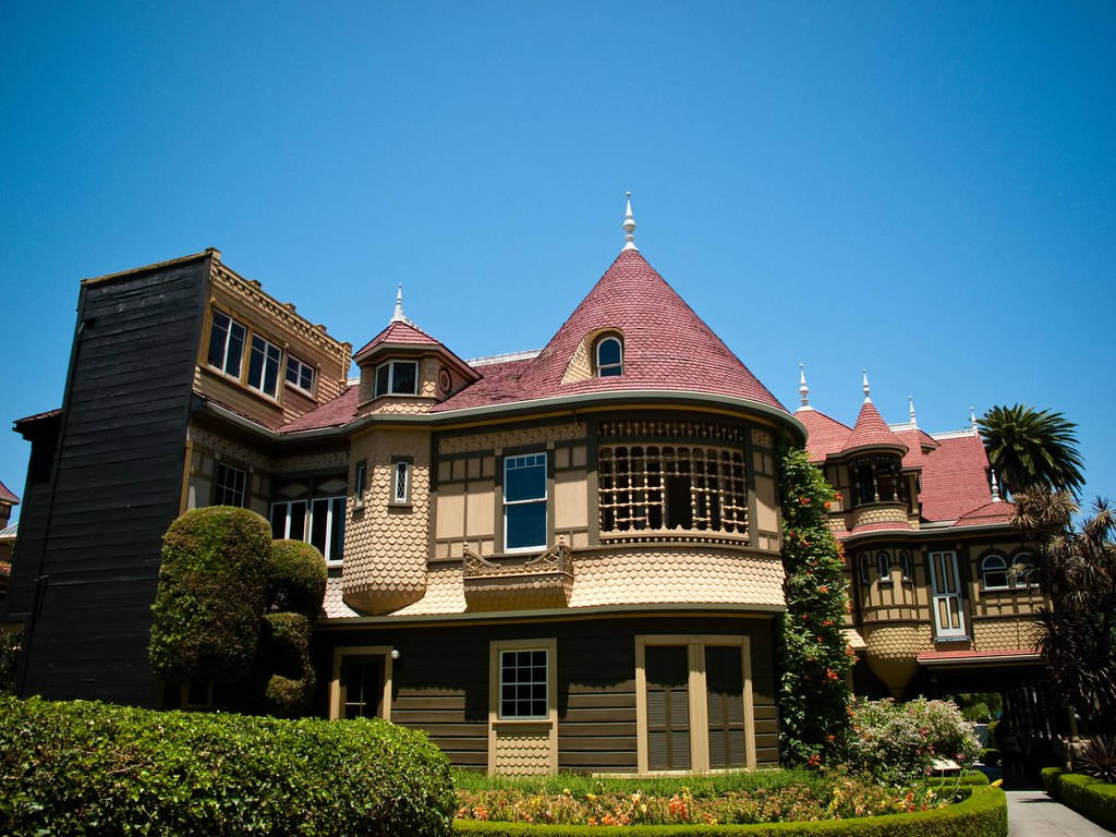 Mansion 桌布 壁紙 — 一座宏偉的維多利亞式豪宅，擁有紅色瓦片屋頂和華麗的木製品，矗立在晴朗的藍天下，周圍環繞著修剪整齊的樹籬和棕櫚樹。