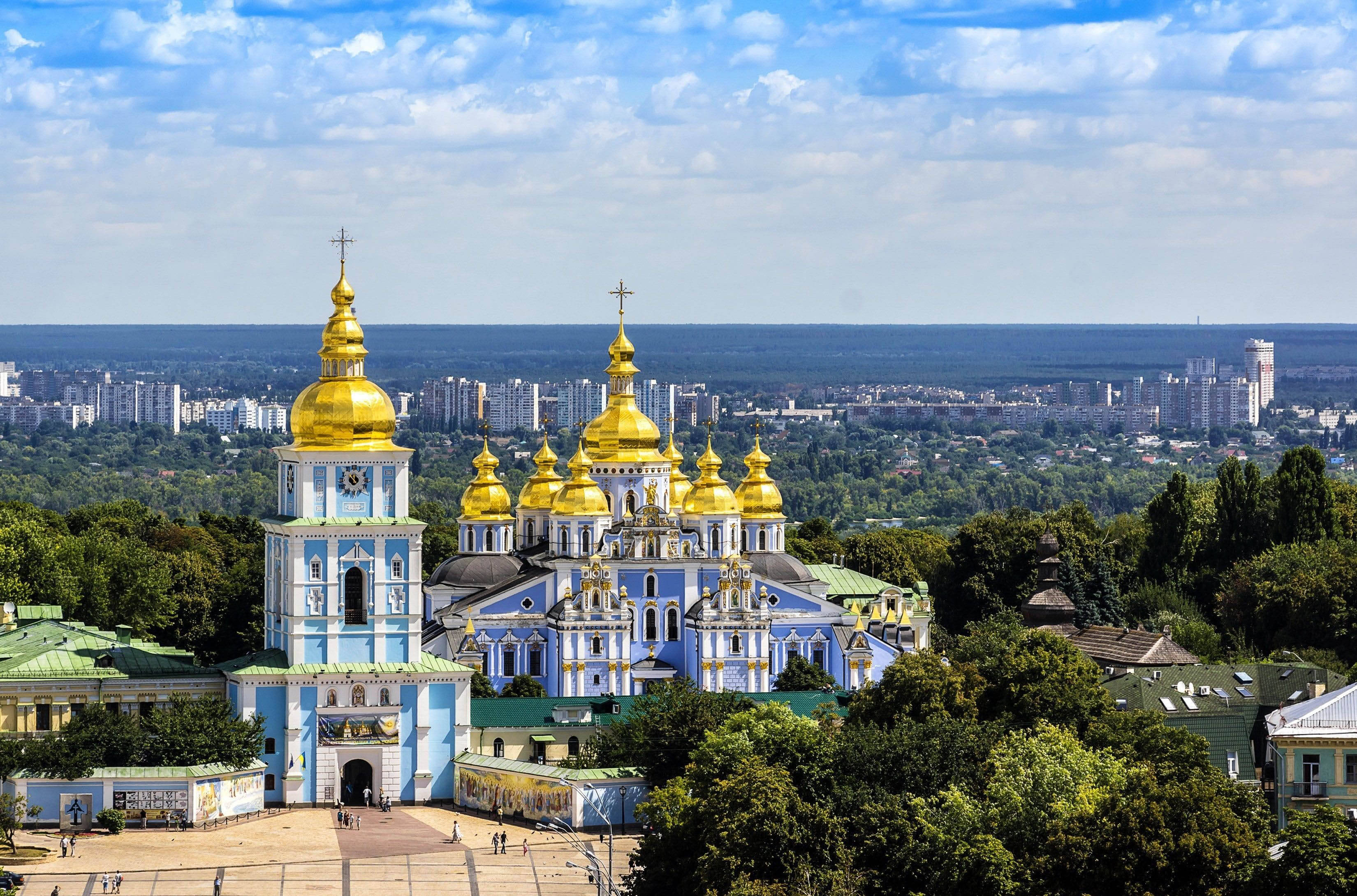 Ukraine 桌布 壁紙 — 一座宏偉的藍色和金色大教堂，有多個圓頂，矗立在茂密的綠樹叢中，俯瞰著廣闊的城市景觀，天空湛藍，白雲點點。