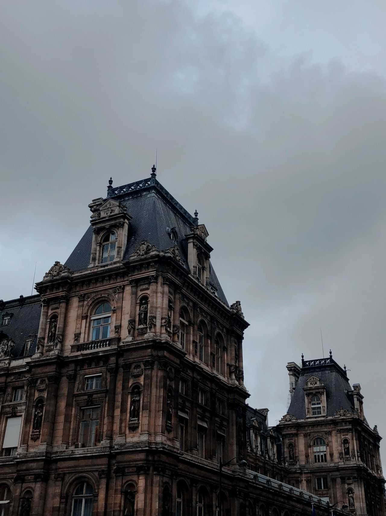 Dark Academia Aesthetic 桌布 壁紙 — 一座宏偉的裝飾性建築，擁有複雜的石雕和深色石板屋頂，矗立在陰沉多雲的天空下。