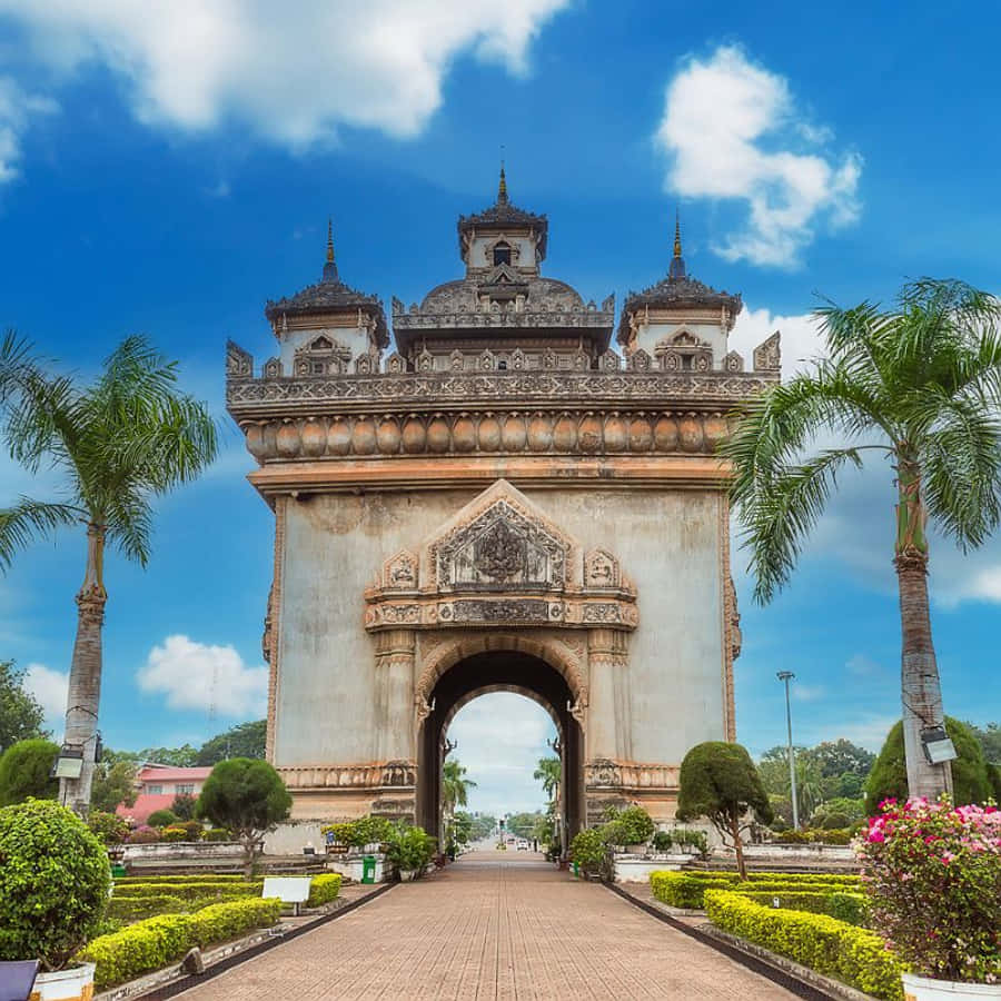 Laos 桌布 壁紙 — 一座宏偉的裝飾性石拱門，帶有精緻雕刻，矗立在明亮的藍天下，兩旁是棕櫚樹和修剪整齊的花園。