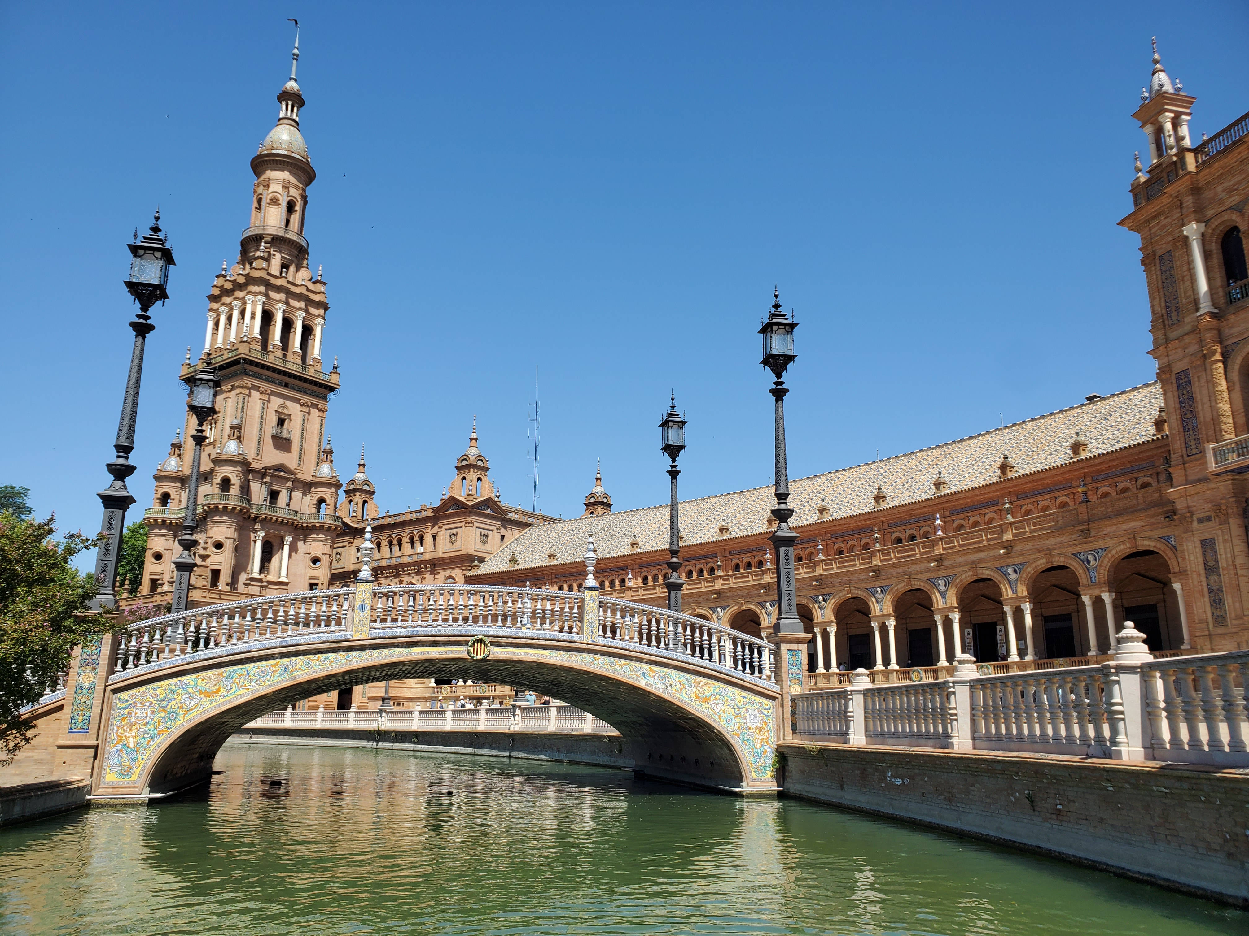 Seville 桌布 壁紙 — 一座宏偉的裝飾橋樑，帶有色彩繽紛的瓷磚拱門，橫跨在清澈藍天下的寧靜運河上，周圍是歷史悠久的西班牙建築。