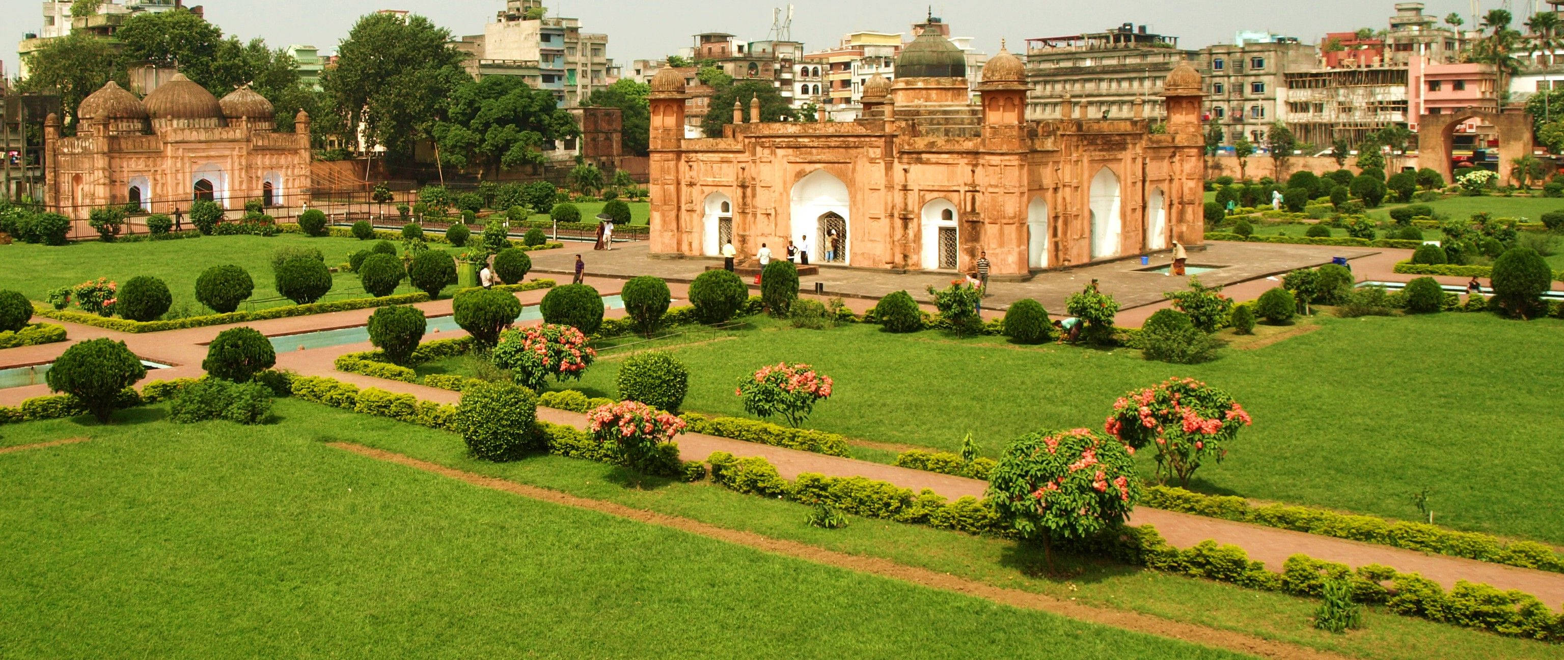 Dhaka 桌布 壁紙 — 一座宏偉的赤陶宮殿，帶有拱形大門，矗立在修剪整齊的花園中，綠草如茵，粉紅色的開花灌木在明亮的天空下。