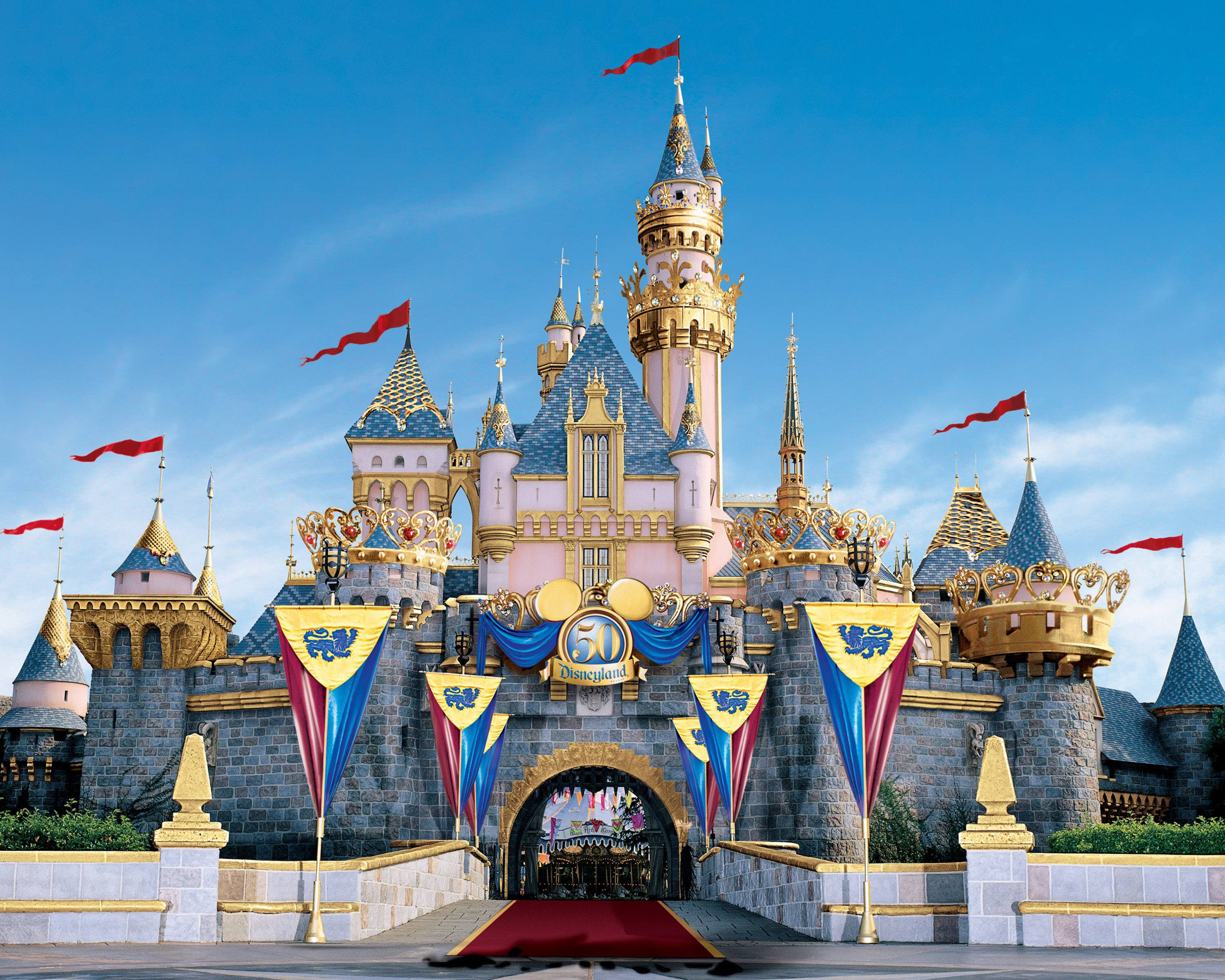 Disneyland Castle 桌布 壁紙 — 一座宏偉的迪士尼城堡，擁有粉紅色的塔樓、金色點綴和紅色旗幟，矗立在清澈的藍天下，紅地毯通往入口。