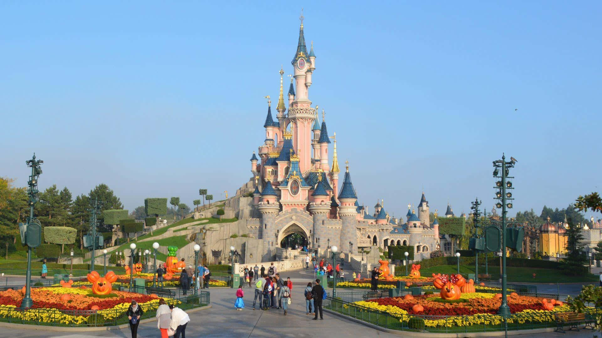 Disneyland Castle 桌布 壁紙 — 一座宏偉的迪士尼城堡，擁有粉紅色的塔樓和藍色的屋頂，在晴朗的藍天下高高聳立，周圍環繞著色彩繽紛的花壇和遊客。