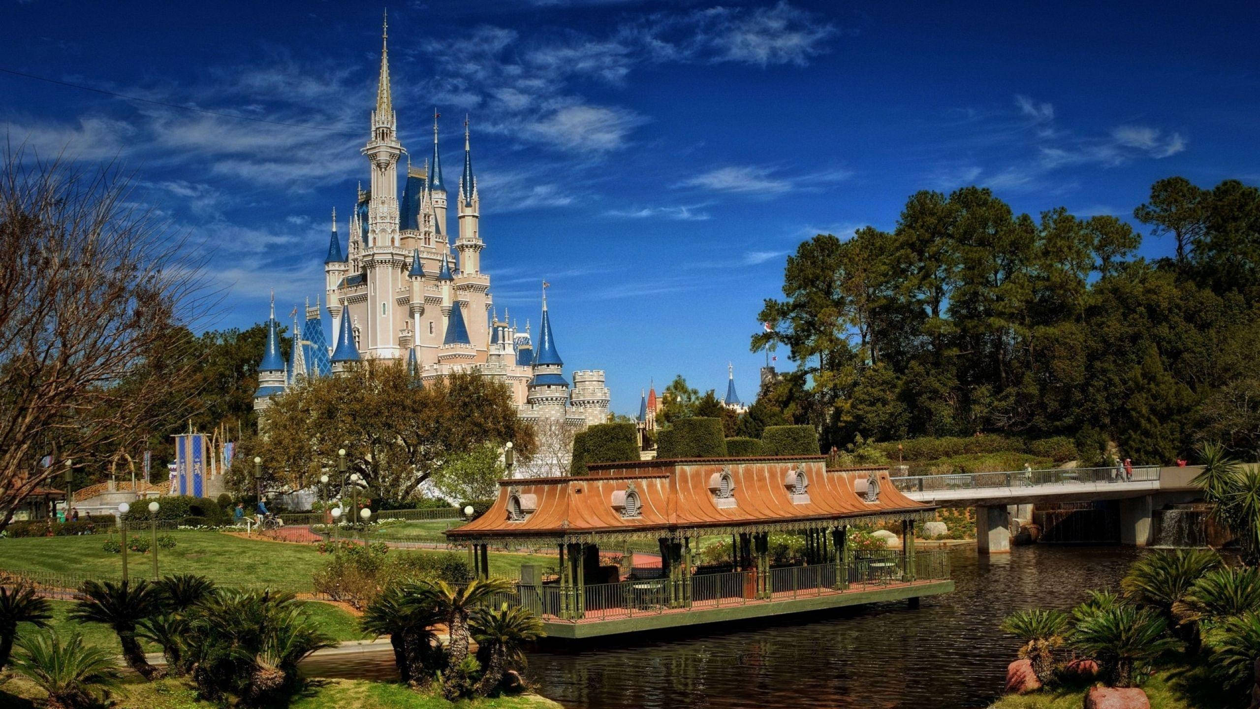 Disneyland Castle 桌布 壁紙 — 一座宏偉的迪士尼城堡，擁有藍色尖頂，在明亮的藍天下，矗立在寧靜的運河和茂密的綠植之後。