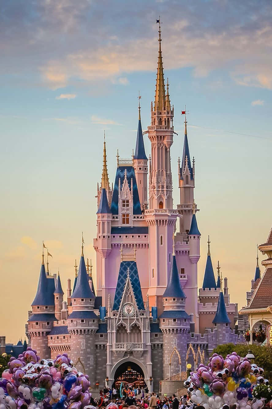 Disney Castle 桌布 壁紙 — 一座宏偉的迪士尼城堡，擁有藍色尖頂和金色點綴，在柔和的日落天空下高高聳立，周圍環繞著人群和彩色氣球。
