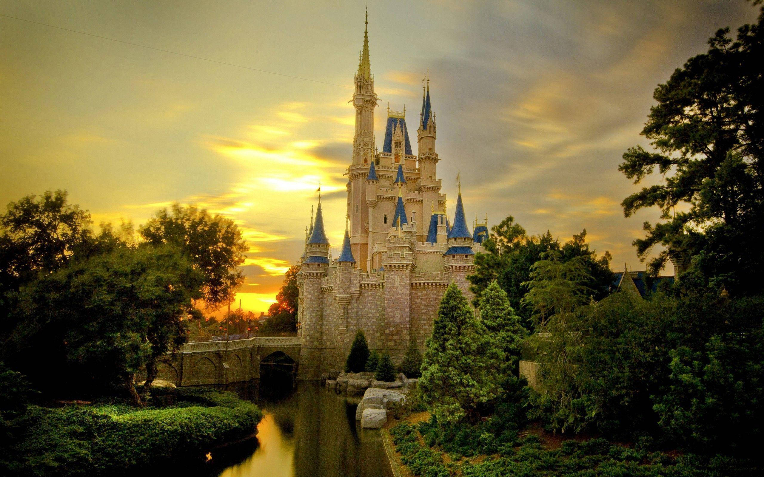 Disneyland 桌布 壁紙 — 一座宏偉的迪士尼城堡，擁有藍色尖塔，矗立在日落時分，倒映在平靜的護城河中，周圍環繞著茂密的綠樹。