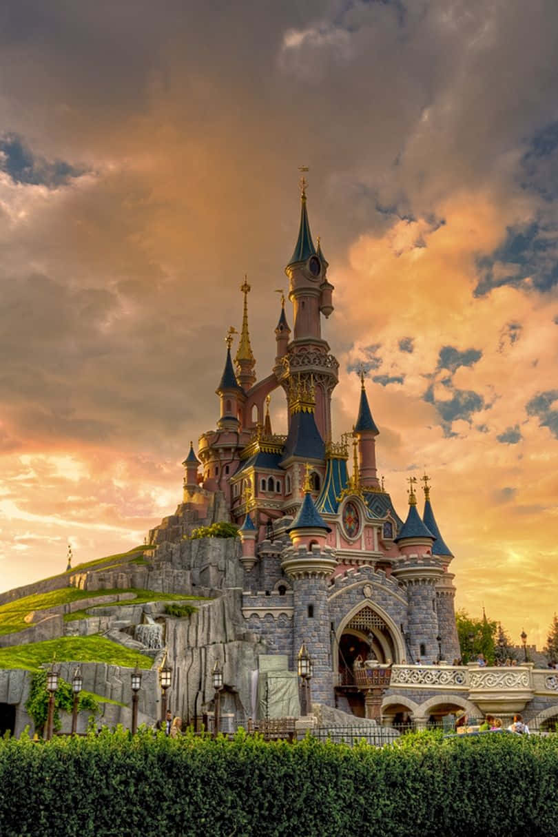 Disneyland Castle 桌布 壁紙 — 一座宏偉的迪士尼城堡，擁有藍色尖頂和粉紅色塔樓，矗立在岩石山丘上，背景是戲劇性的金色日落天空。