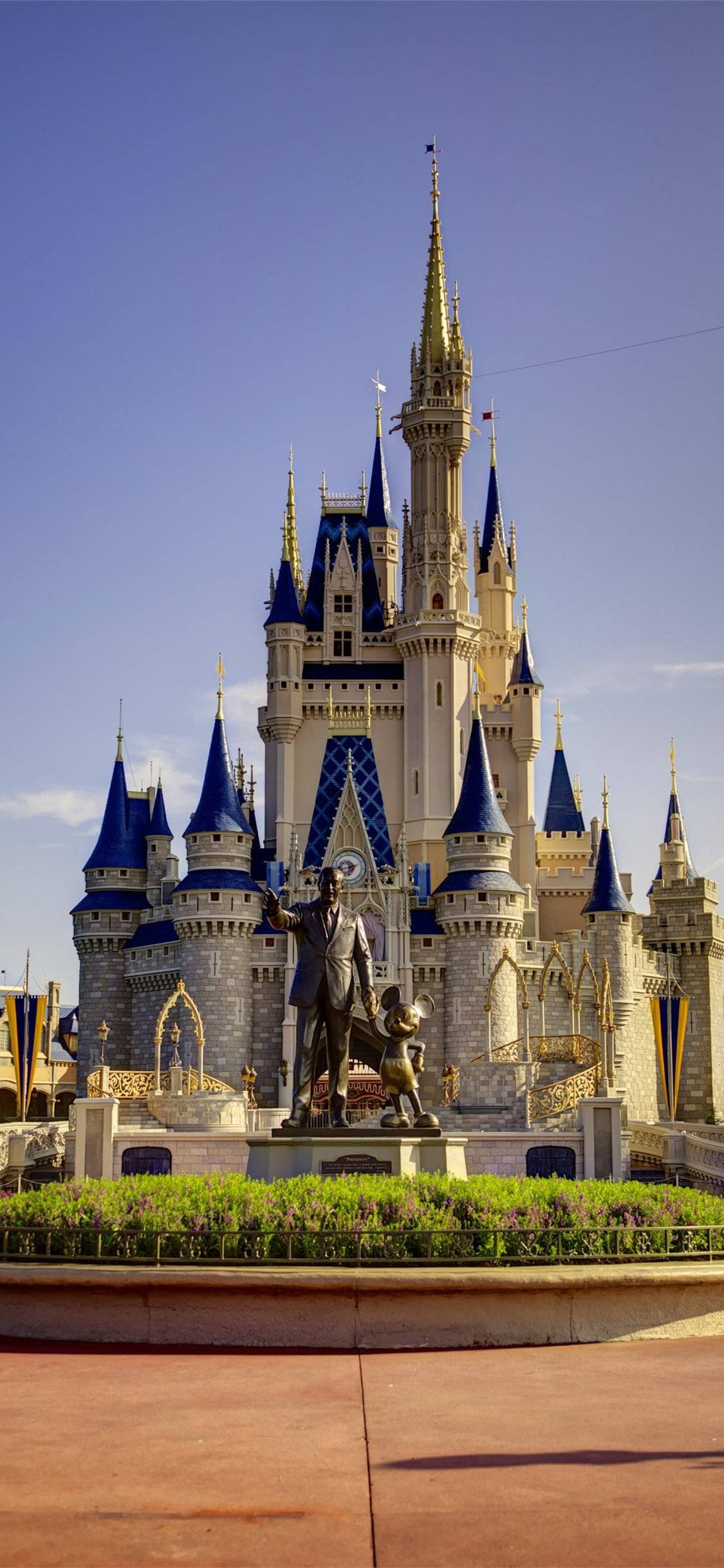 Disneyland 桌布 壁紙 — 一座宏偉的迪士尼城堡，擁有藍色尖頂和金色點綴，在晴朗的藍天下高高聳立，前面有一座雕像。
