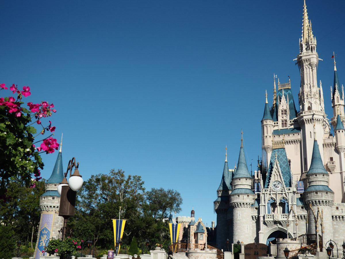 Disney Castle 桌布 壁紙 — 一座宏偉的迪士尼城堡，擁有藍色尖頂，在藍天白雲下高聳入雲，周圍環繞著粉紅花朵和茂密綠植。