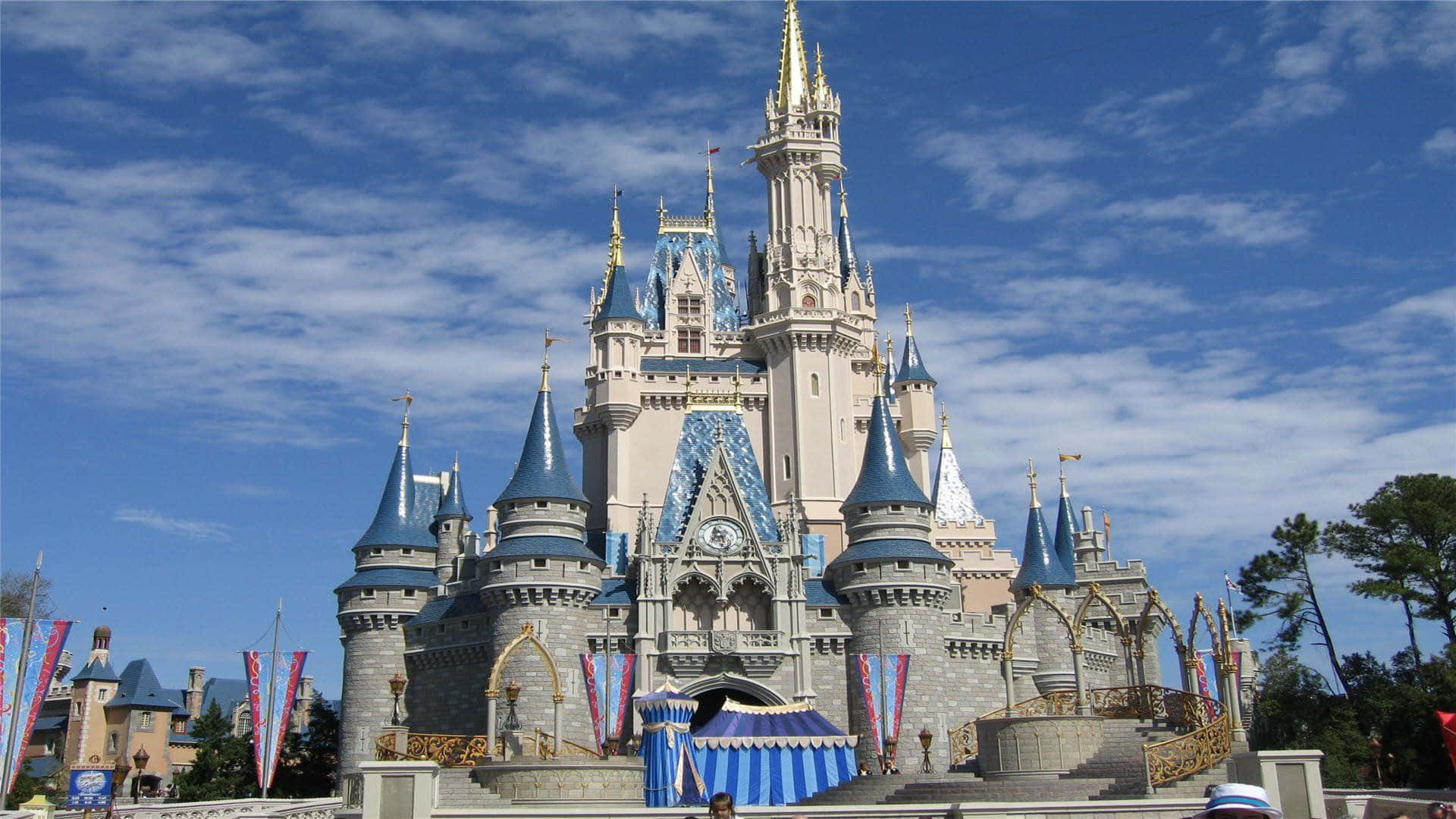 Disney Castle 桌布 壁紙 — 一座宏偉的迪士尼城堡，擁有藍色尖頂和金色點綴，在藍天白雲下巍然聳立。