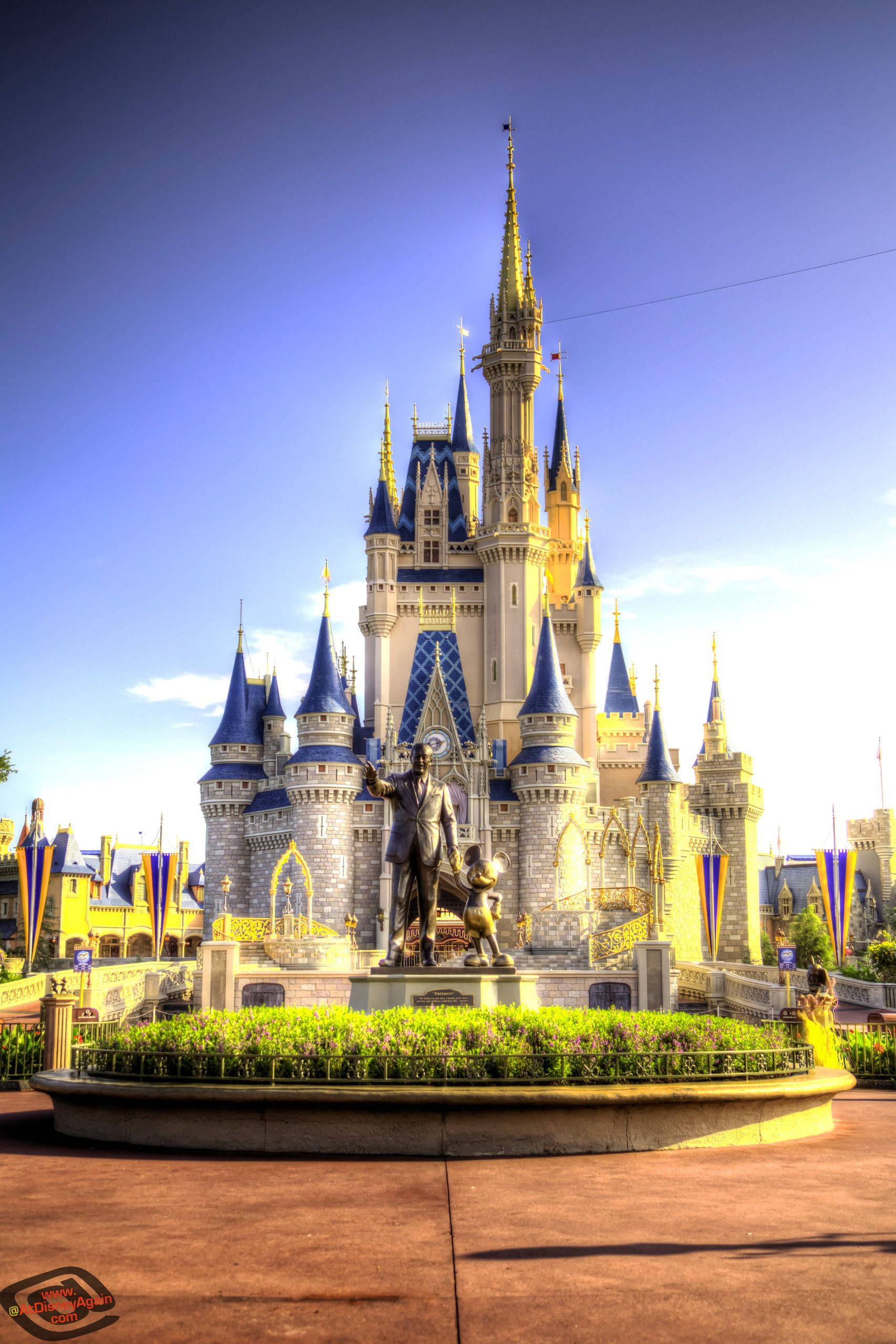 Disney Castle 桌布 壁紙 — 一座宏偉的迪士尼城堡，擁有藍色屋頂和金色尖塔，在晴朗的藍天下巍然聳立，前面有一座雕像。