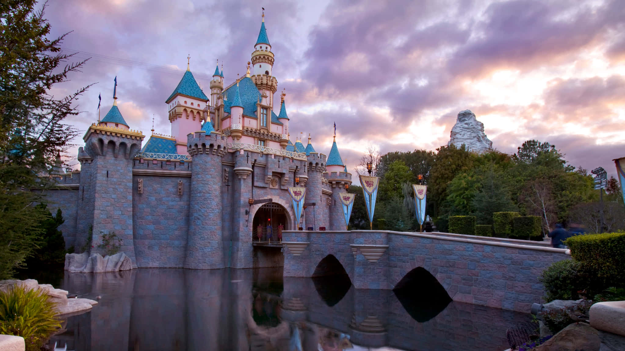 Disneyland 桌布 壁紙 — 一座宏偉的迪士尼城堡，擁有藍色屋頂和石塔，在暮色天空下倒映在平靜的水面上，天空中有粉紅色和紫色的雲彩。