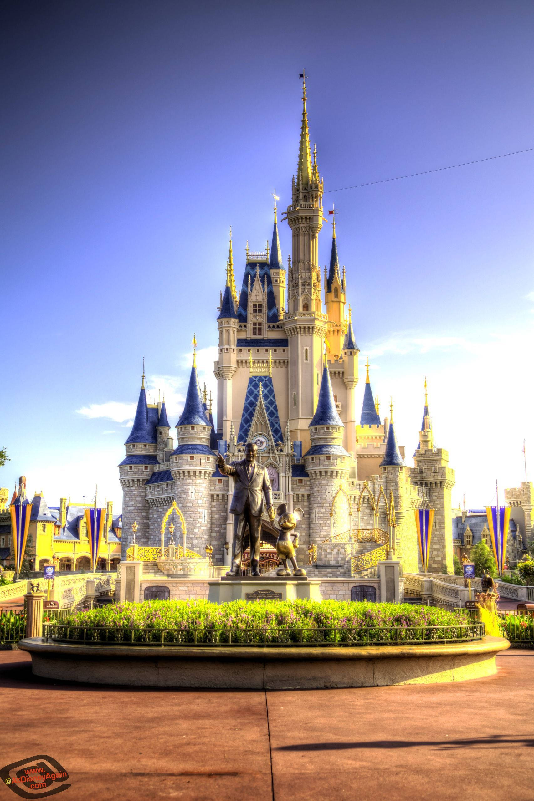 Disneyland Castle 桌布 壁紙 — 一座宏偉的迪士尼城堡，擁有藍色屋頂和金色尖塔，在晴朗的藍天下高高聳立，前面有一座雕像。