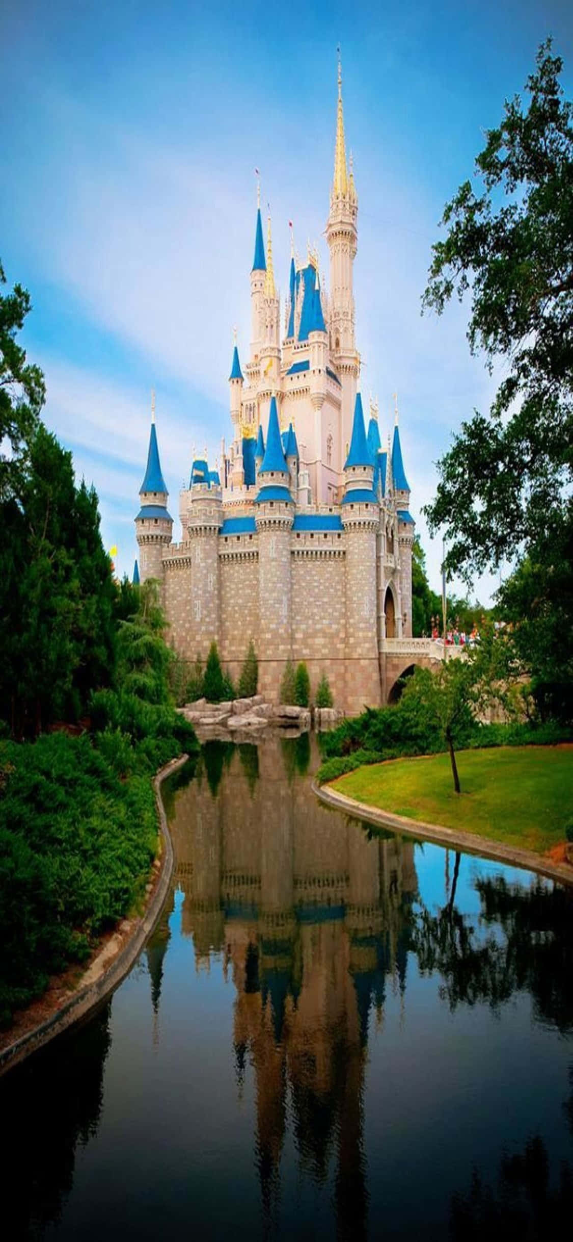 walt disney 桌布 壁紙 — 一座宏偉的迪士尼城堡，藍色尖頂完美倒映在平靜的運河中，四周環繞著茂密的綠樹，天空清澈湛藍。