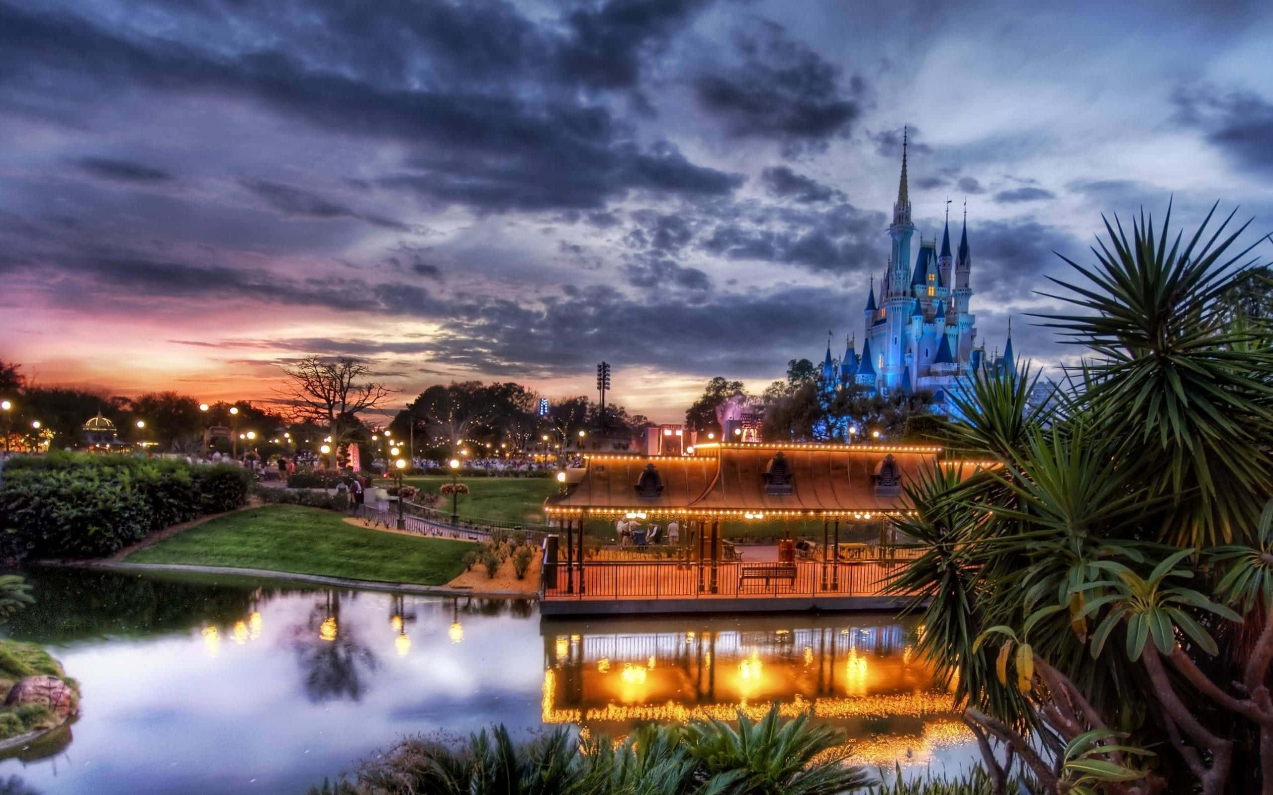 Disney World Desktop 桌布 壁紙 — 一座宏偉的迪士尼城堡在暮色天空下發出藍光，燈光倒映在平靜的湖面上，四周環繞著茂密的綠植和棕櫚樹。