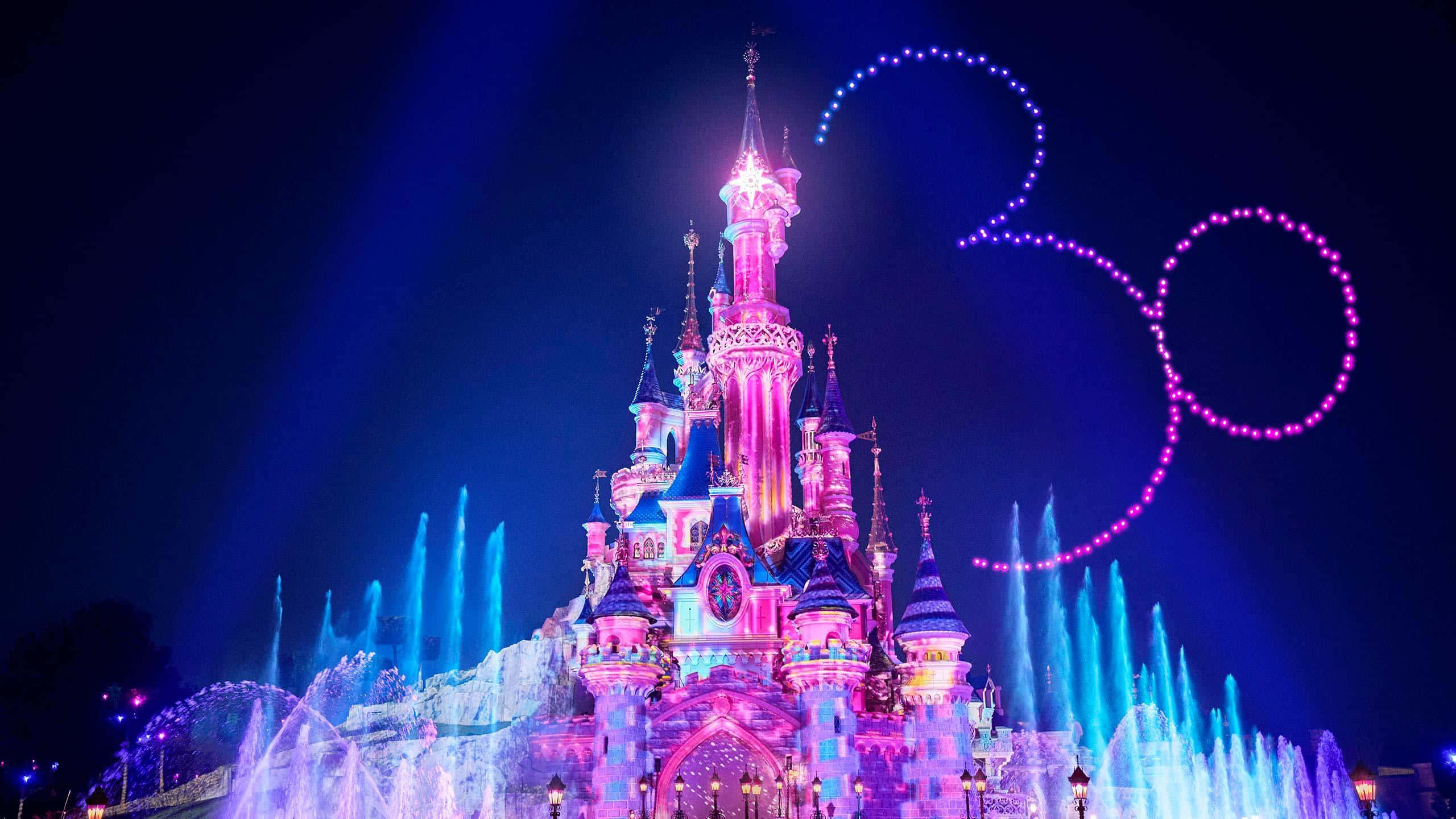Disneyland 桌布 壁紙 — 一座宏偉的迪士尼城堡在粉紅色和紫色燈光下閃耀，帶有發光的米奇老鼠剪影和夜晚充滿活力的噴泉。